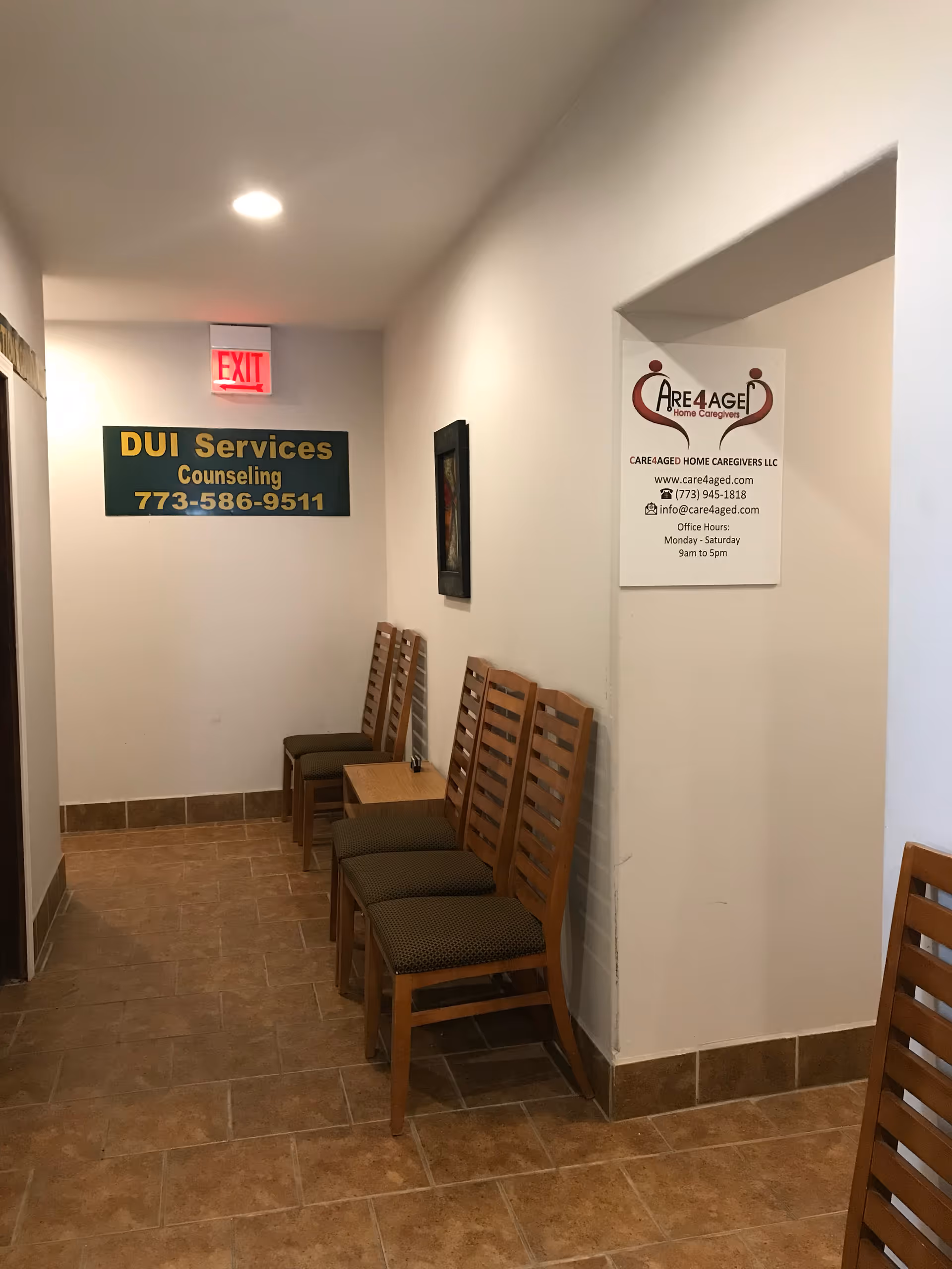 Indoor hallway with wooden chairs along the wall, small table, and exit sign.
