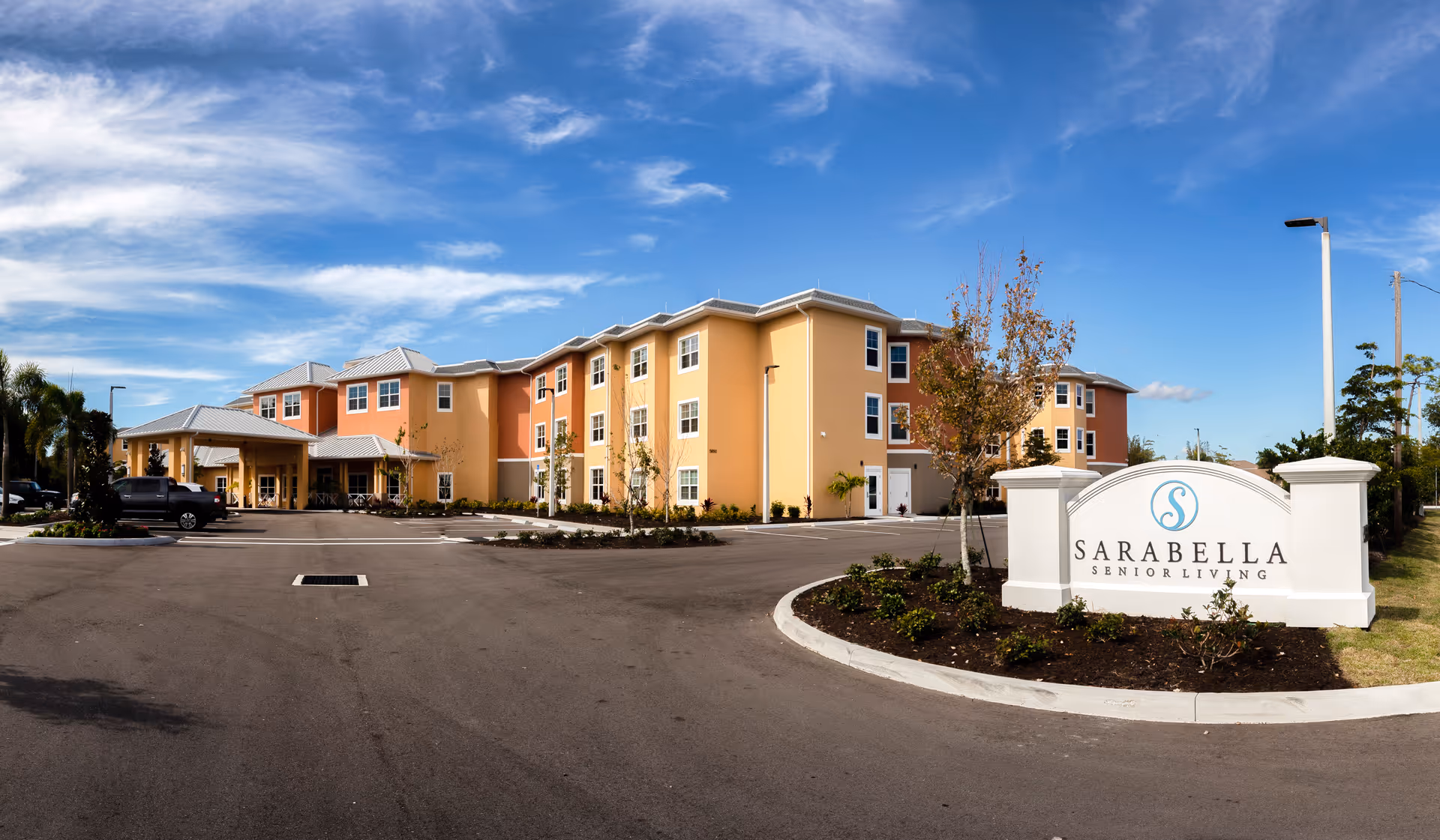 Exterior view of SaraBella Senior Living facility showing a large, three-story building with a covered entrance, surrounded by a parking lot and landscaped areas under a blue sky with scattered clouds.