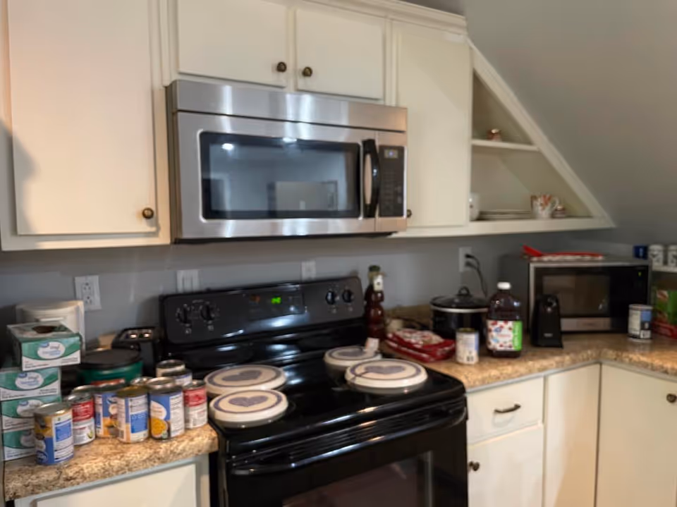 Compact kitchen with white cabinets, microwave, stove, and canned goods on counter.