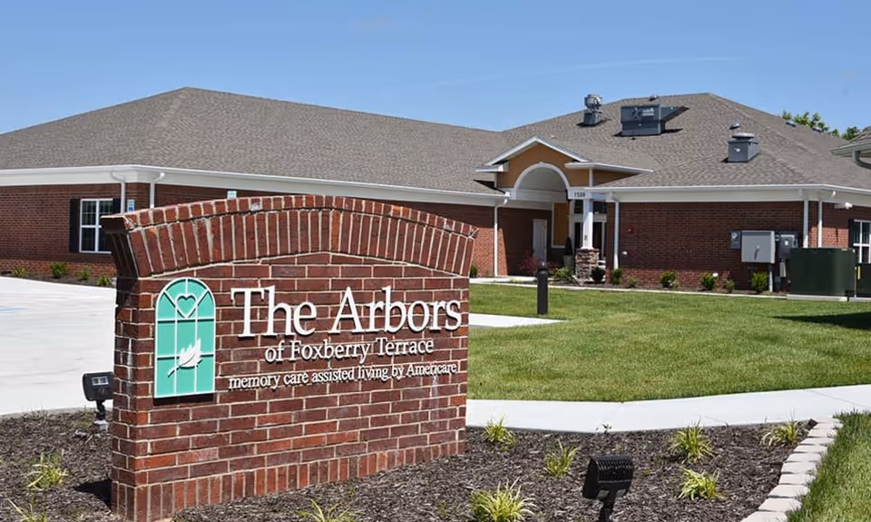Brick entrance sign reading "The Arbors of Foxberry Terrace" in front of a one-story red-brick senior living building and lawn.