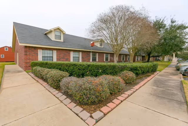 Exterior view of a single-story brick building with multiple windows, surrounded by trimmed bushes and trees. A concrete sidewalk runs alongside the building and a parking area is visible on the right side.