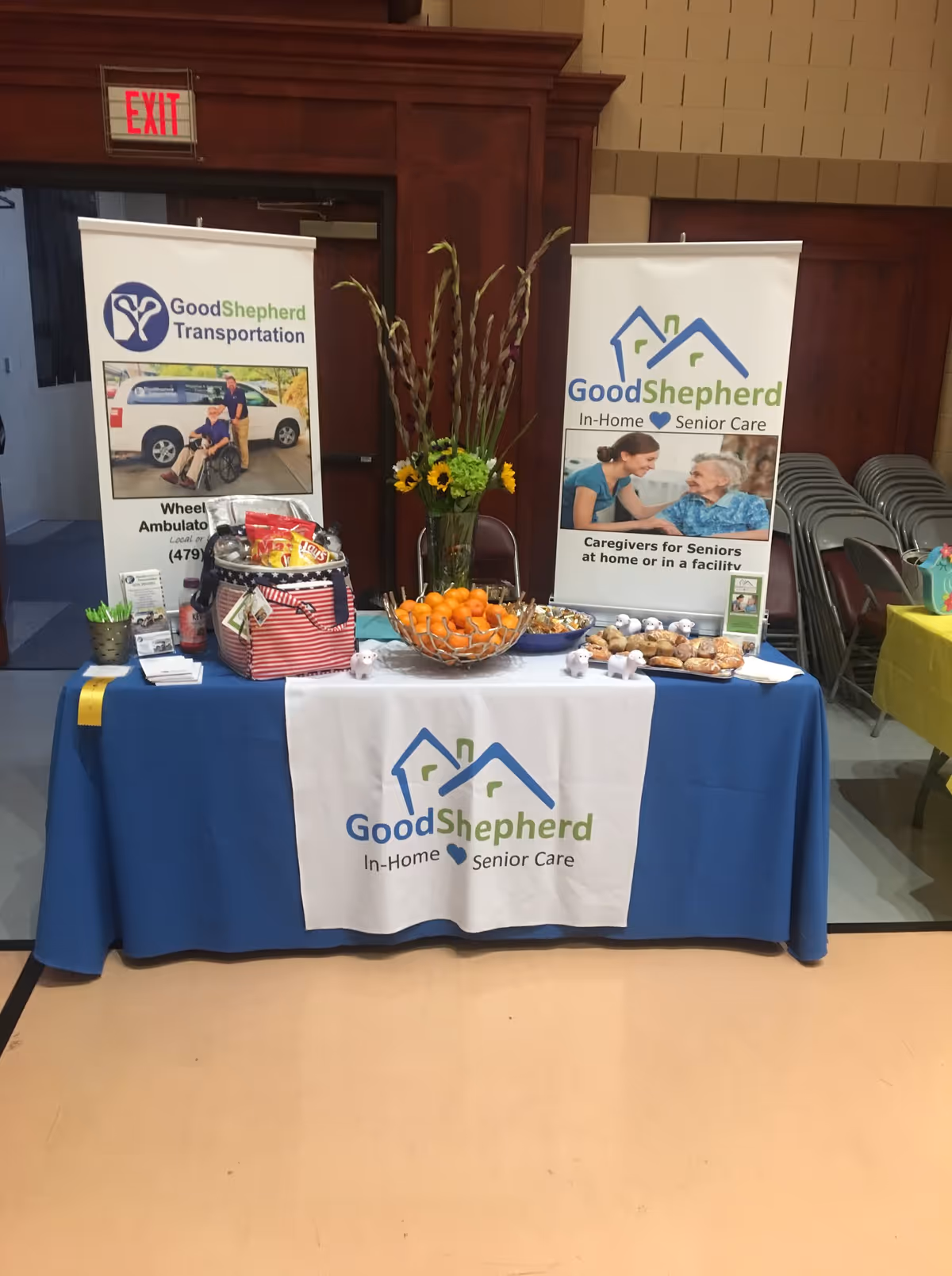 Promotional display table with Good Shepherd banners and fruit centerpiece inside a facility.