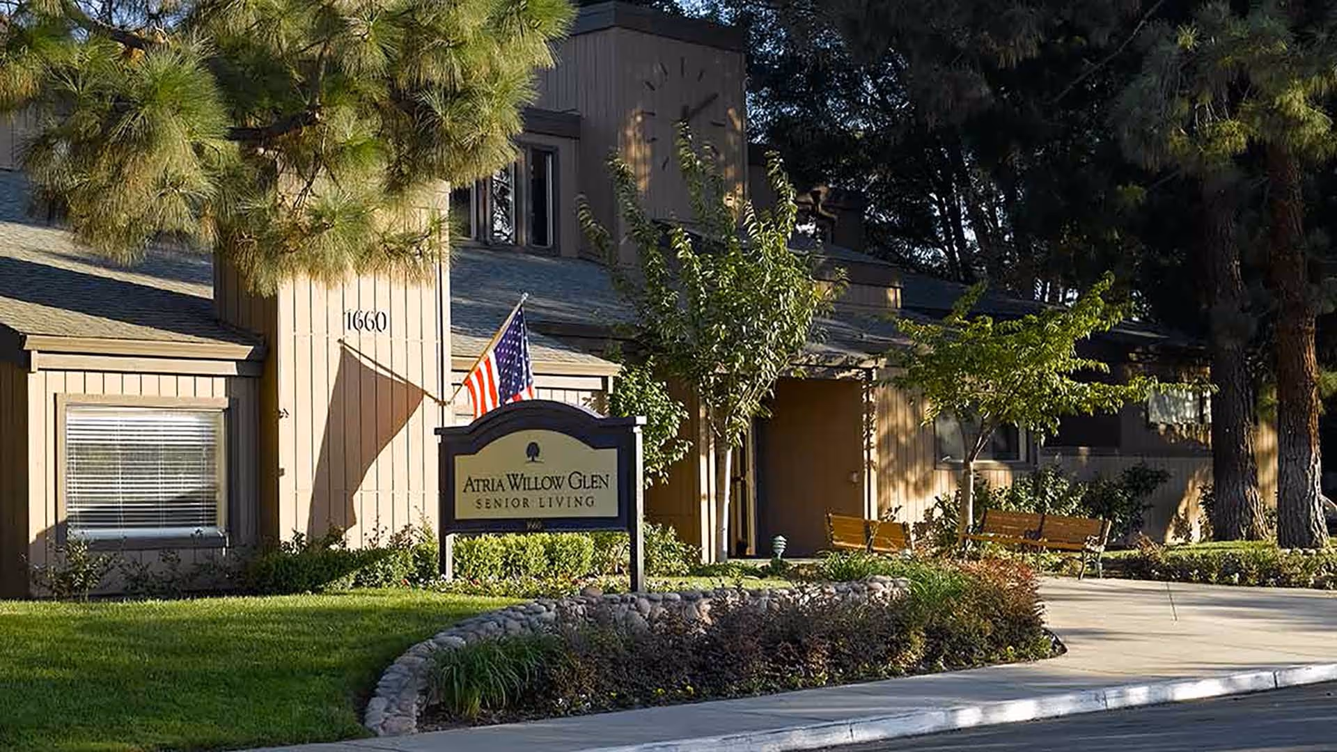 Front entrance of Atria Willow Glen senior living facility showing the building facade, a sign, an American flag, benches, and landscaped grounds.