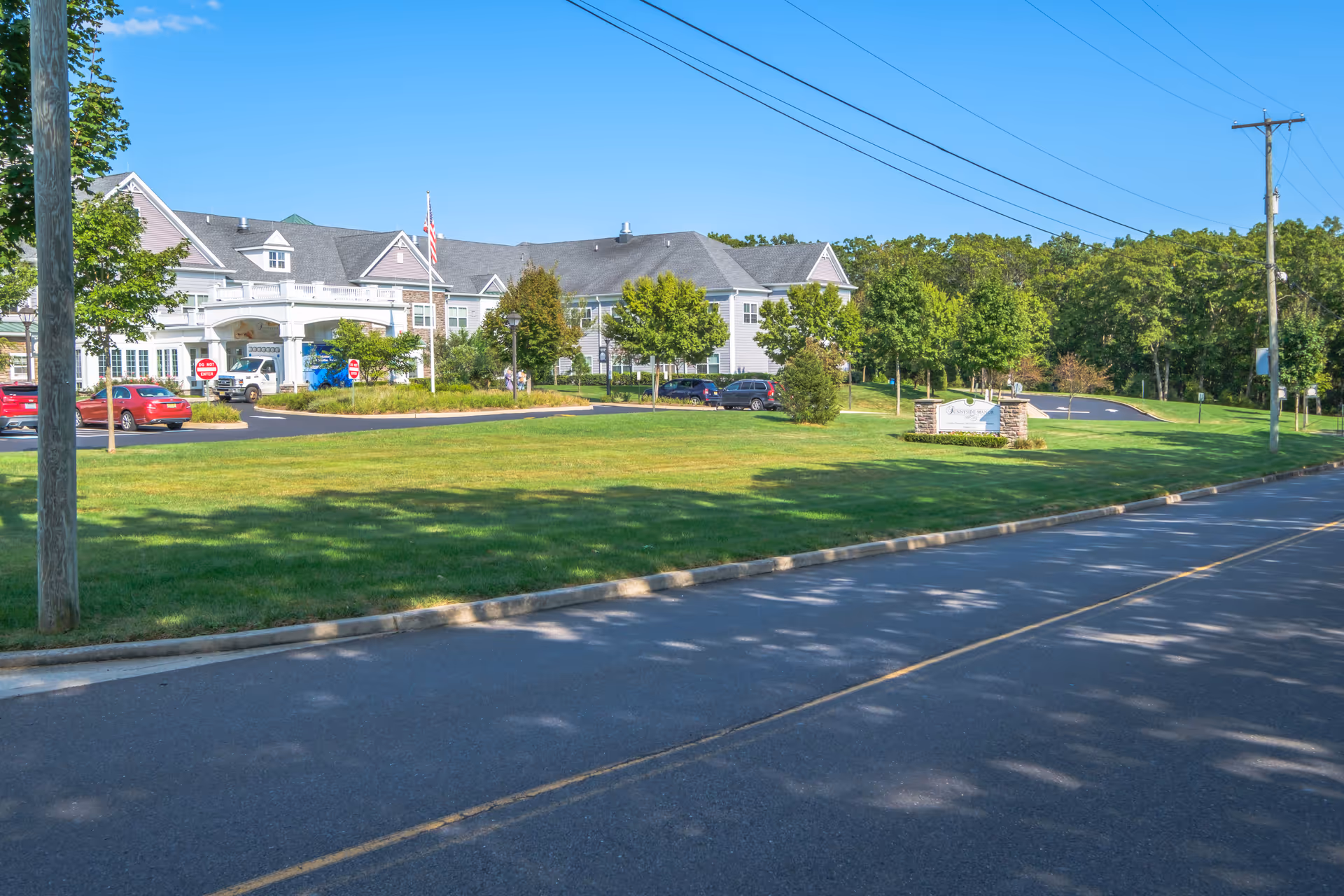 Exterior view of Sunnyside Manor, a large senior living facility with a well-maintained lawn, trees, and a parking area. The building has a gray roof and light-colored siding, with an American flag near the entrance. A sign with the facility's name is visible on the right side near the road.