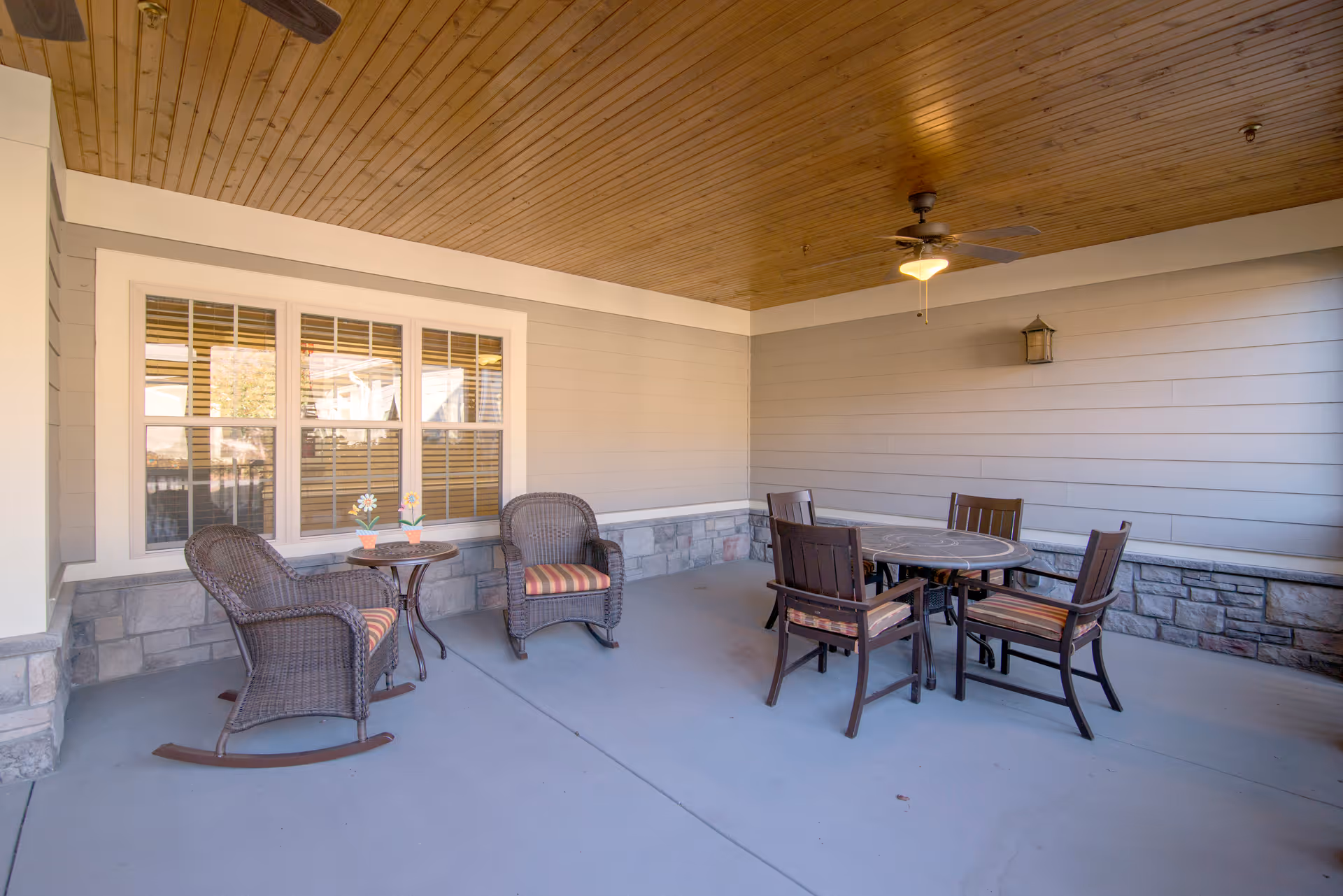 Covered patio with wicker rocking chairs and a round dining table with four chairs under a wood-paneled ceiling and ceiling fan.
