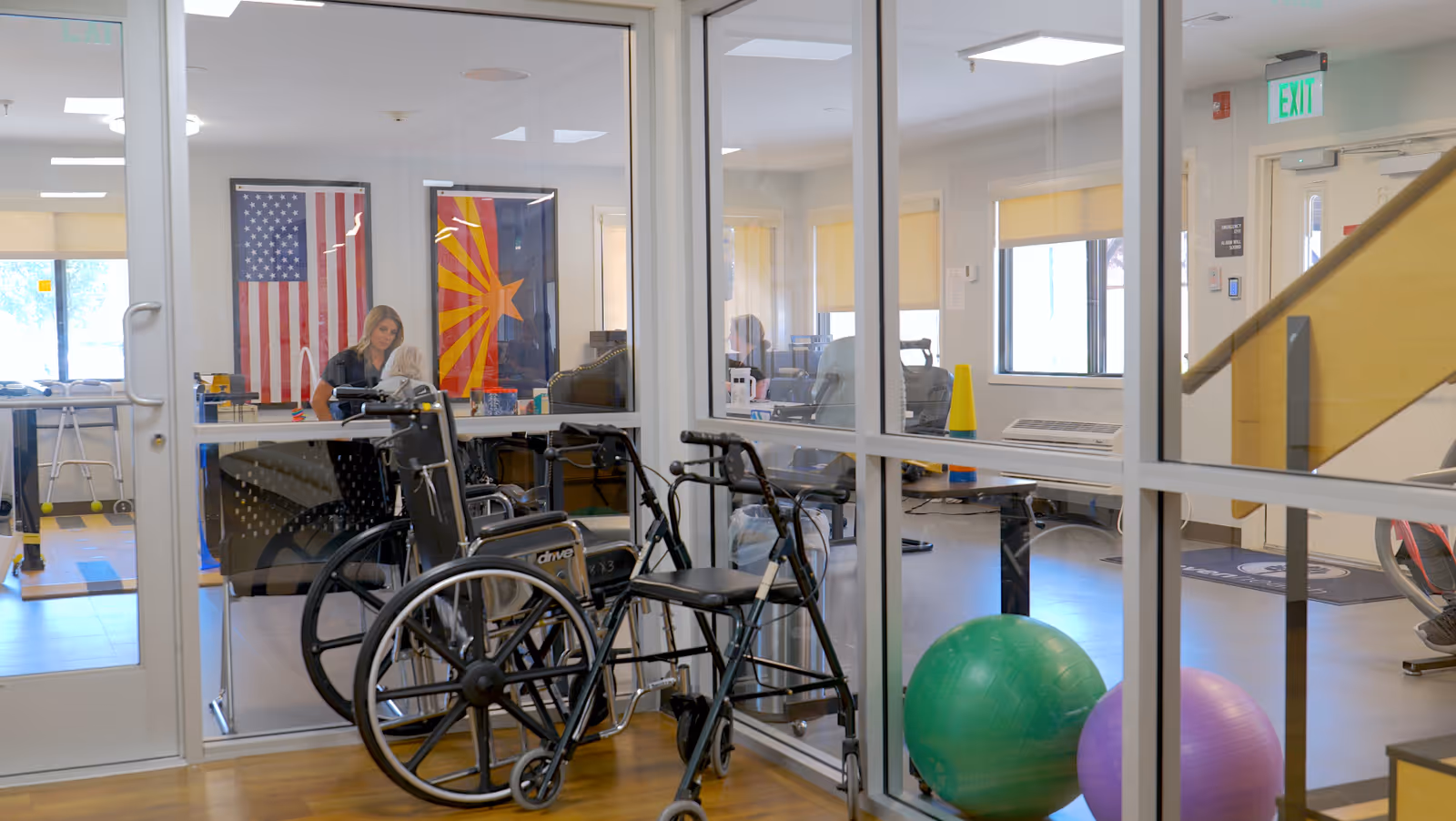 View through glass of a senior facility common area with wheelchairs and walkers in the foreground, exercise balls, and a staff member at a desk with American and Arizona flags on the wall.