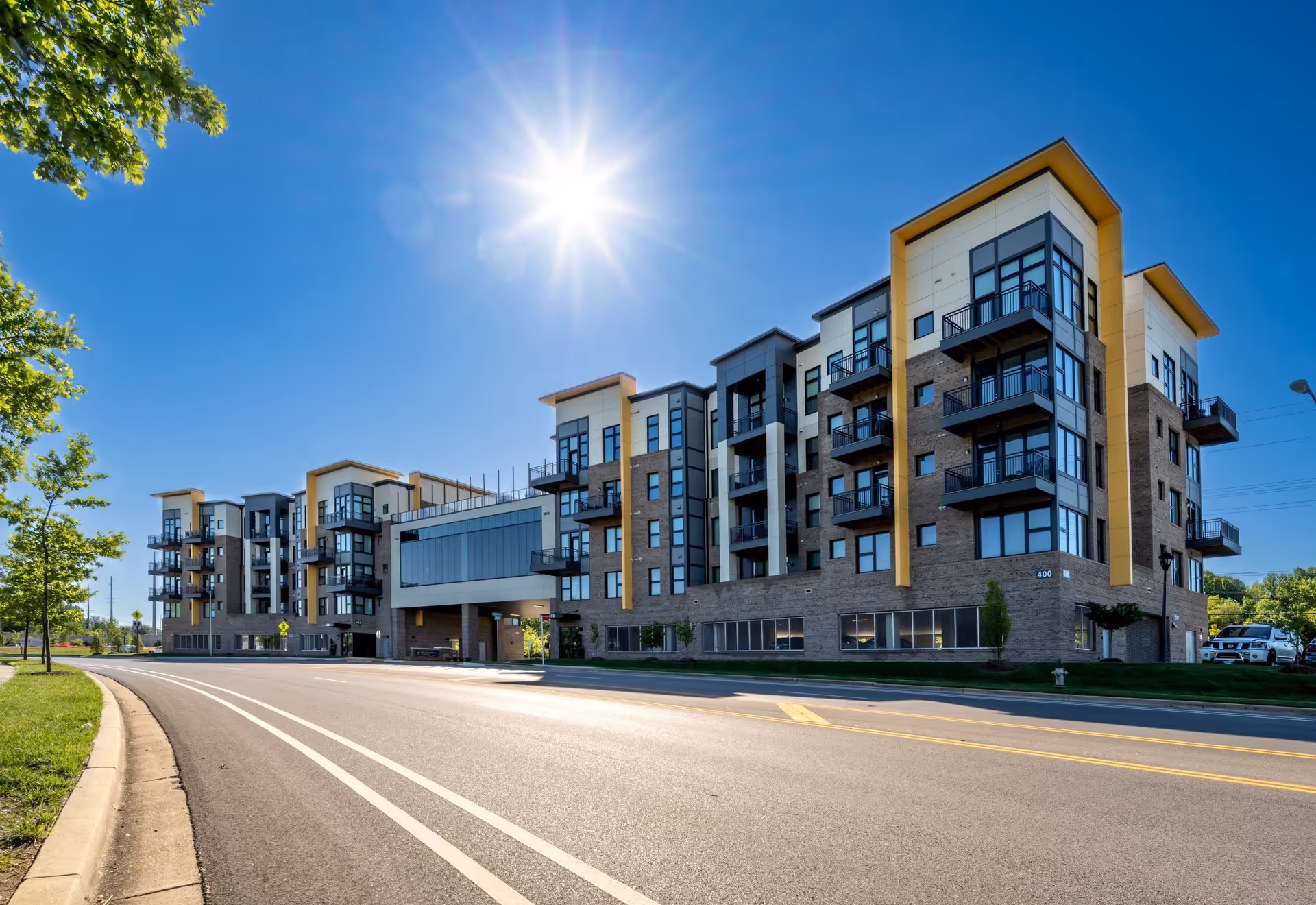 Modern multi-story residential building with balconies and large windows under a bright sunny sky, viewed from across a curved road with green trees on the side.