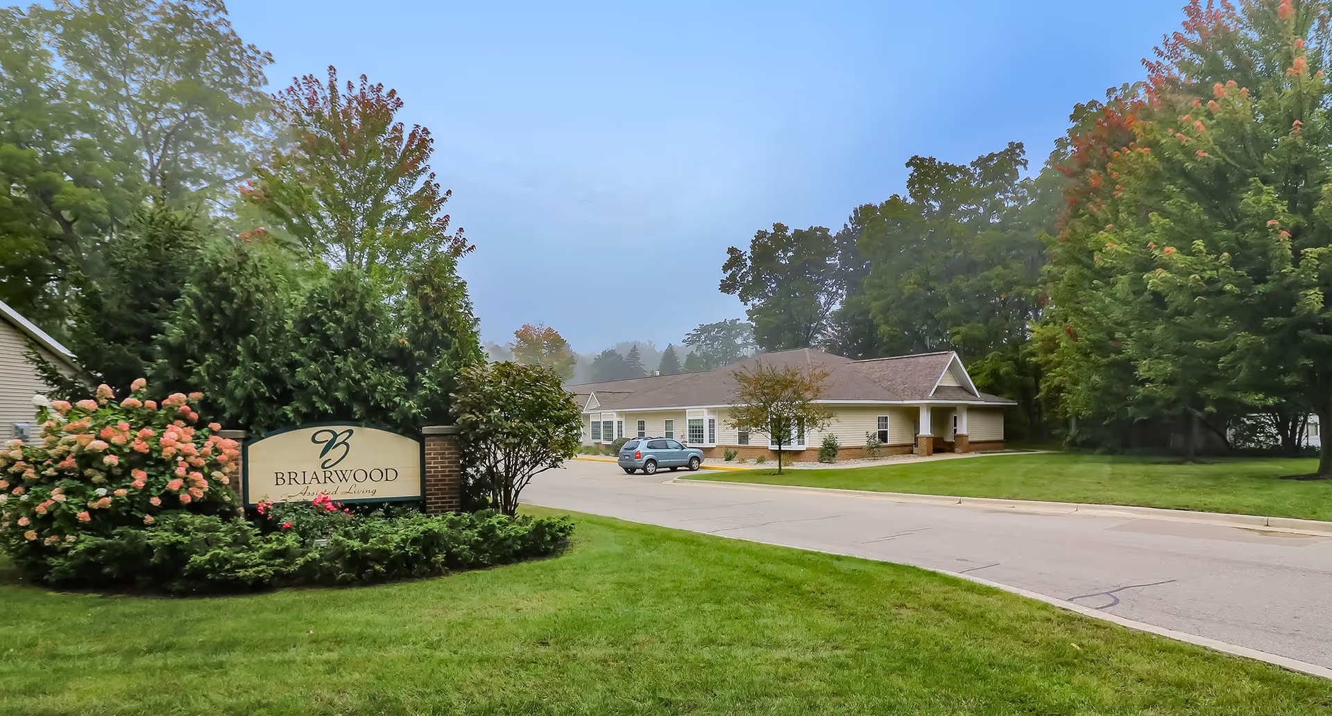 Exterior view of Briarwood Assisted Living & Memory Care facility with a sign surrounded by greenery and flowers on the left, a driveway leading to a single-story building, and trees in the background under a cloudy sky.