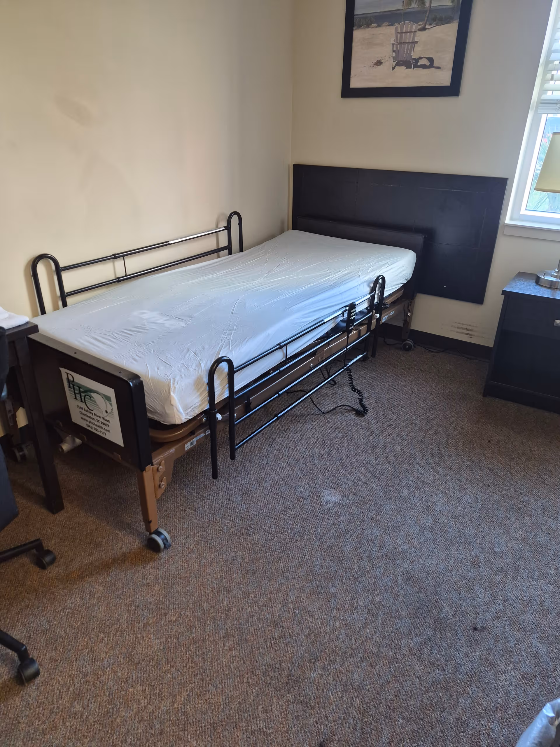 A senior living facility bedroom with a metal-frame hospital bed, headboard, nightstand, and window.