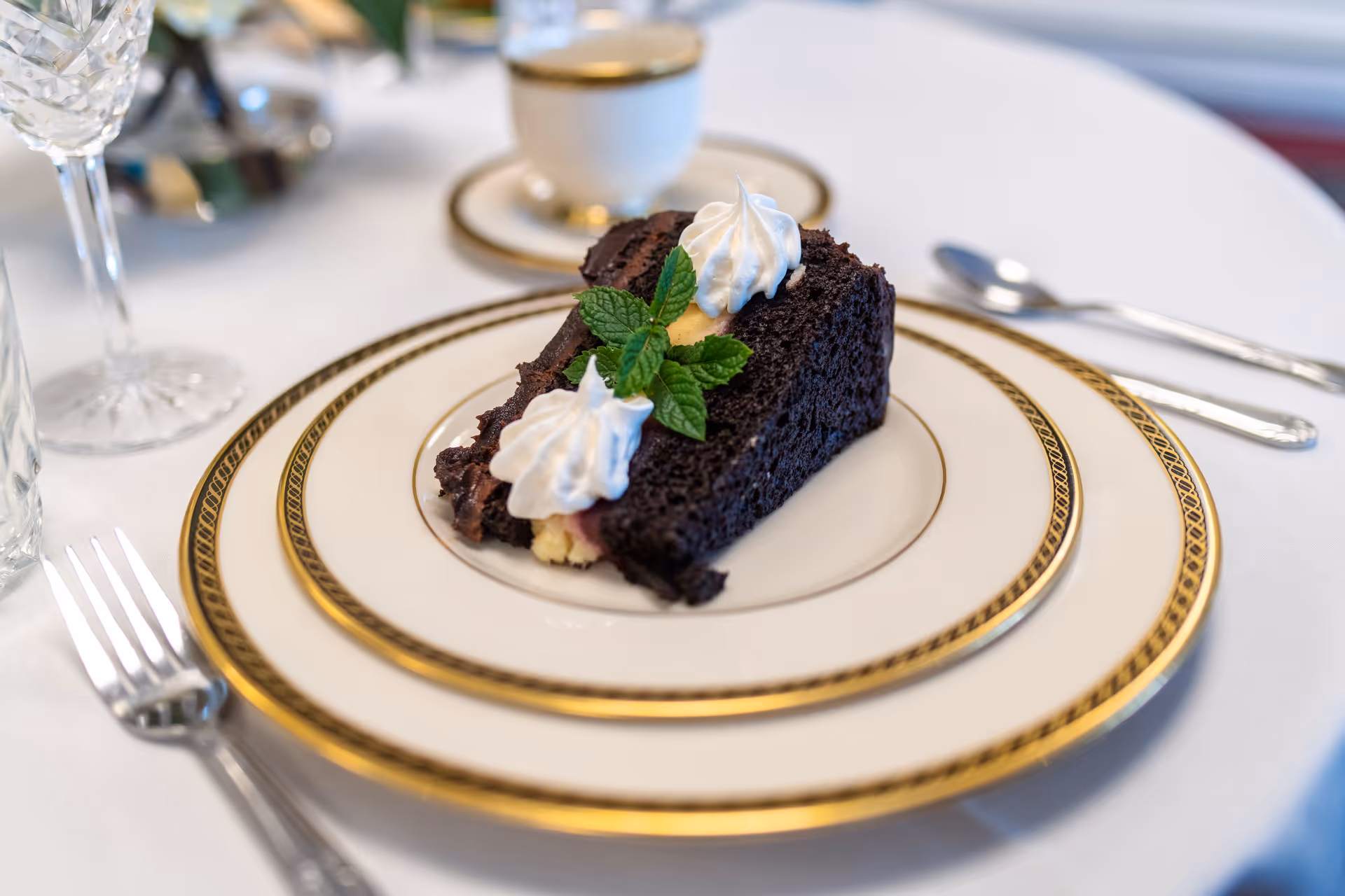 A slice of chocolate cake with white cream and a sprig of mint on top, served on a white plate with gold trim. The plate is on a white tablecloth with a fork, spoon, crystal glass, and a cup and saucer in the background.