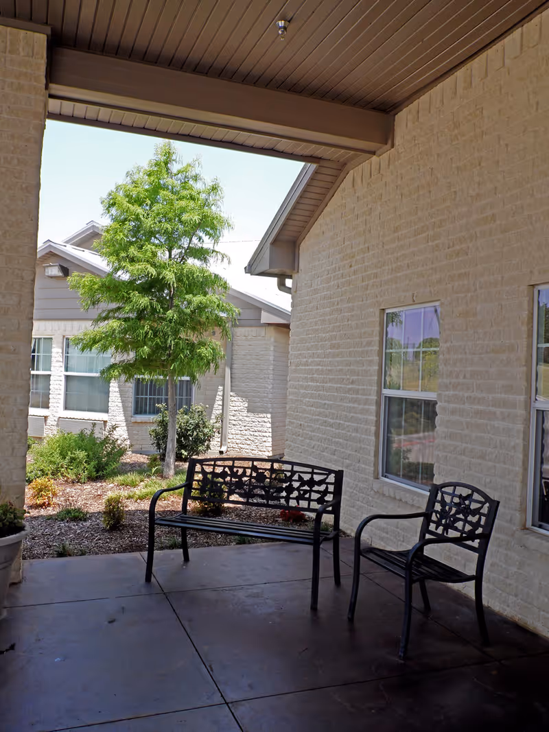 Covered exterior patio/entry with a metal bench and chair beside a light brick building and landscaped yard.