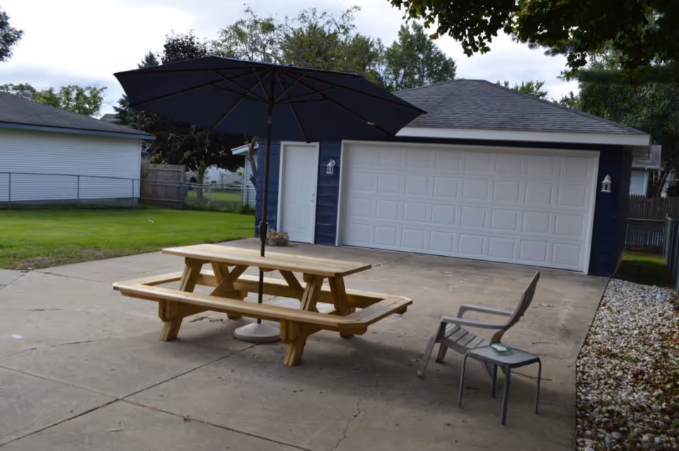 Concrete patio with a wooden picnic table and large black umbrella, a plastic chair and small side table in front of a detached two-car garage.
