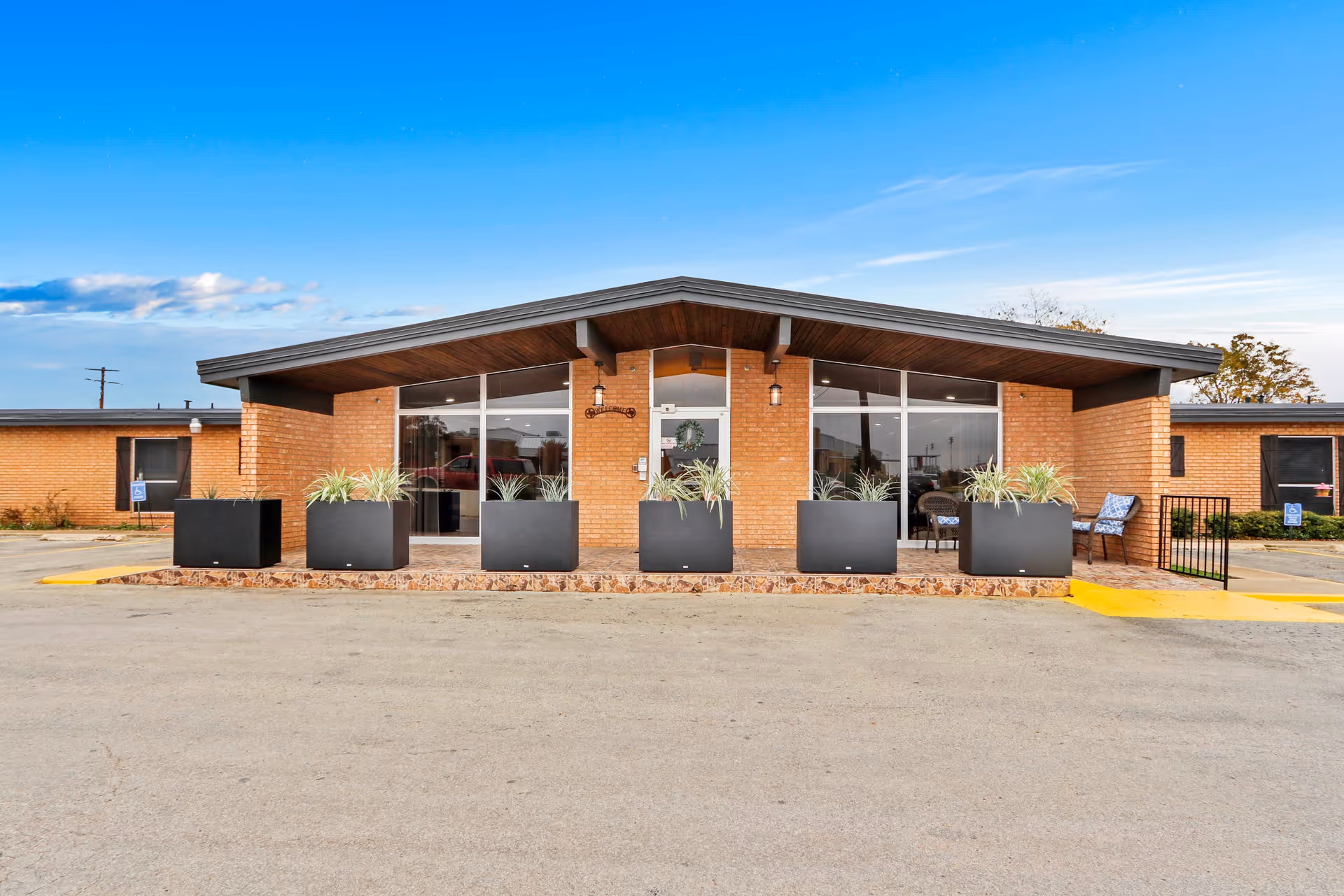 Single-story brick nursing facility front entrance with a wide overhanging roof, large windows, black planters and a paved parking area.