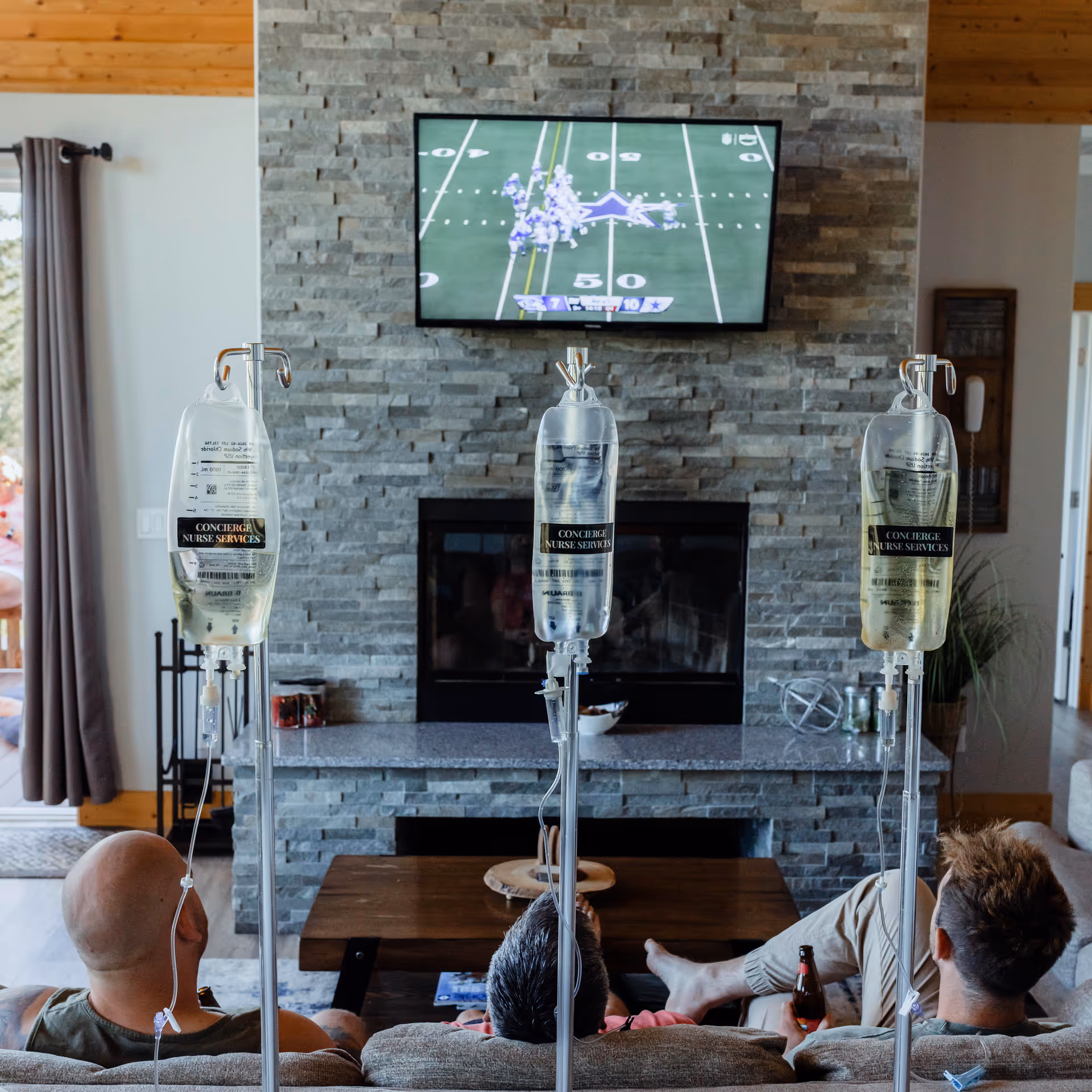 Three IV stands align in a living room, with residents watching TV by a stone fireplace.