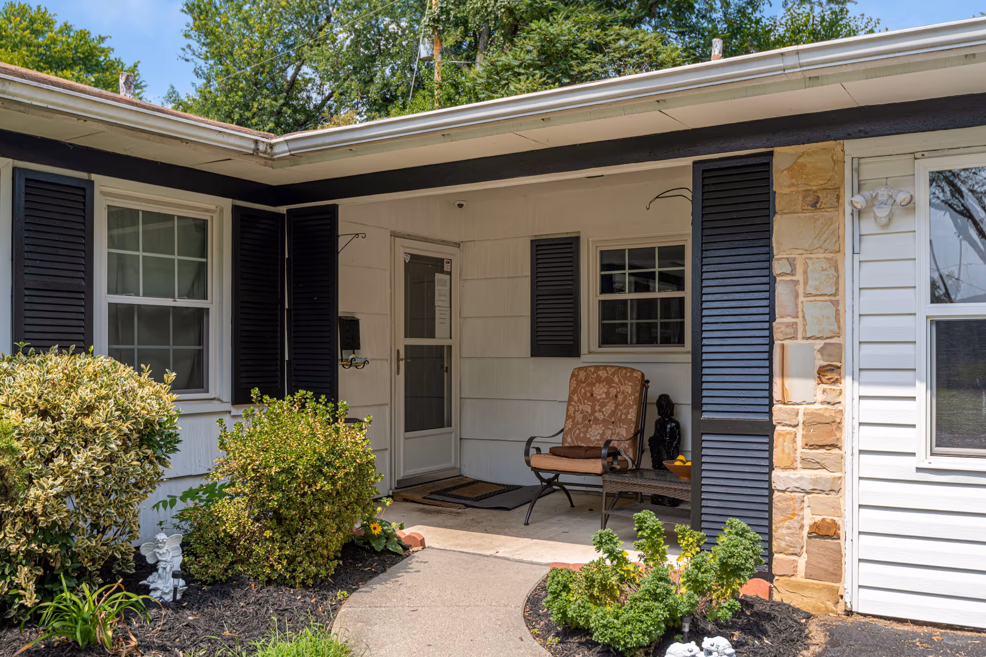 Front porch area of a residential assisted living facility with a cushioned chair, small table, and decorative plants around the entrance. The building exterior features white siding, black shutters, and a stone accent wall.