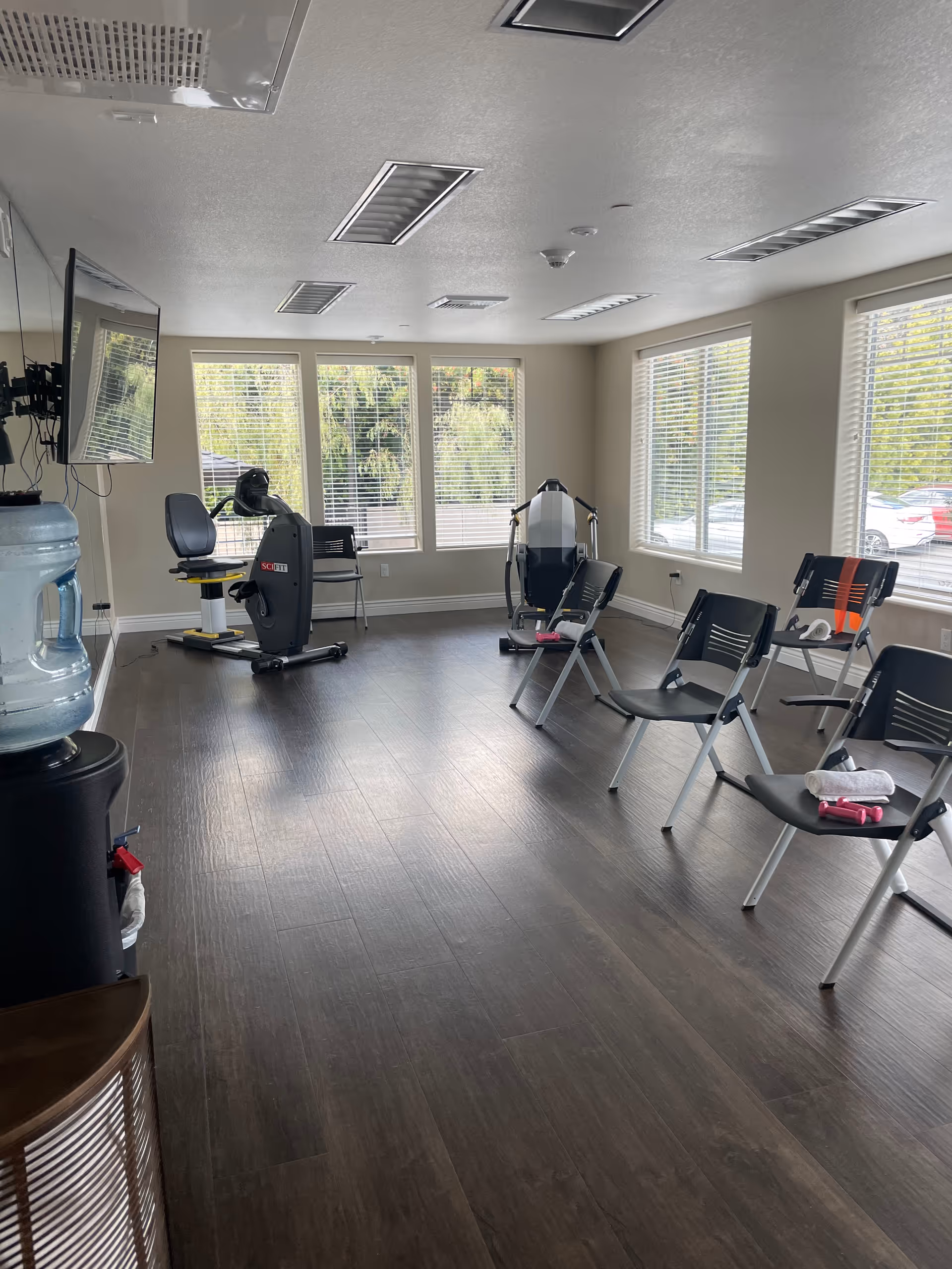 Interior fitness room with exercise machines, chairs, and a water cooler near windows.