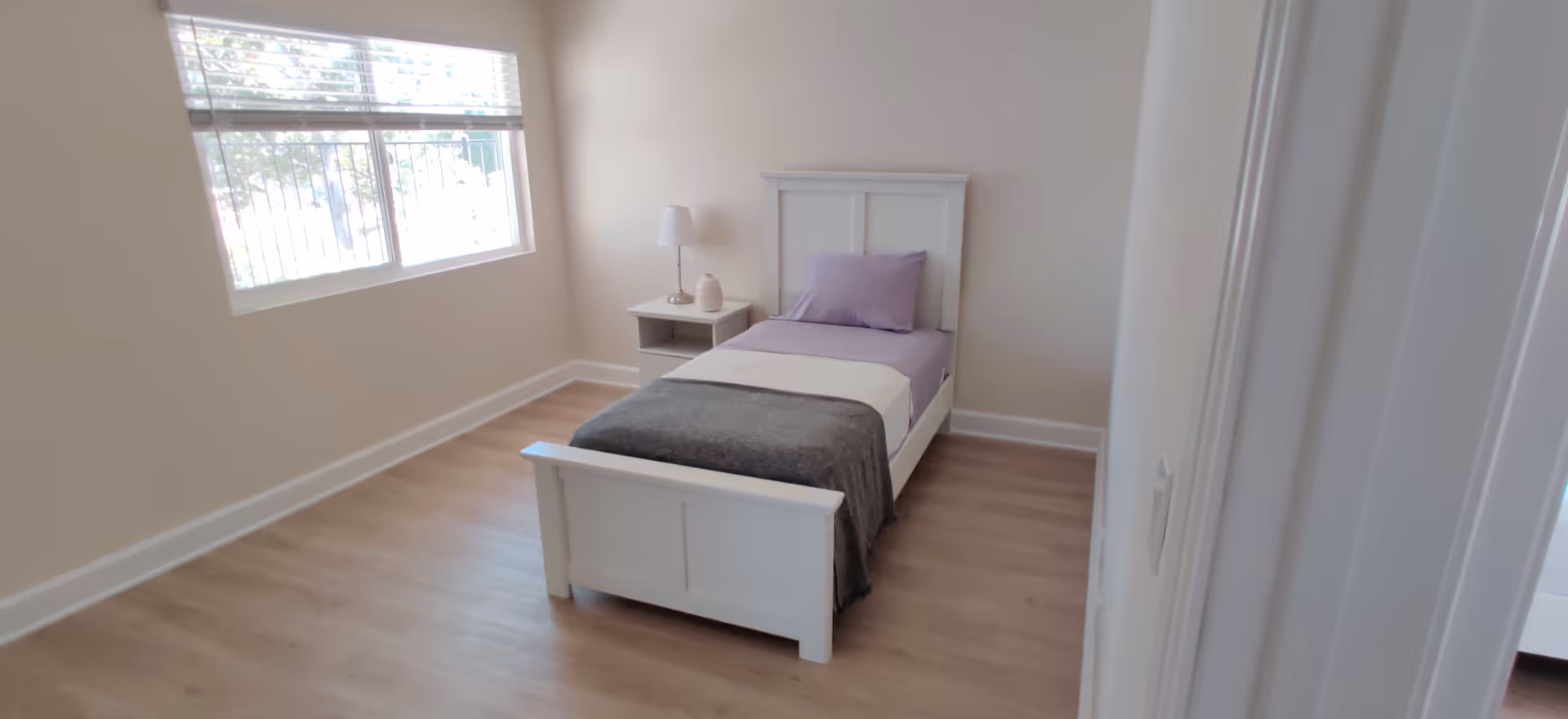 A small, tidy bedroom with a white bed, lavender bedding, and a nightstand.