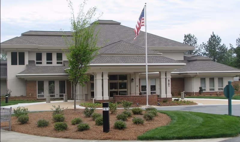Front entrance of a community building with a covered porte-cochere, American flag, and landscaped circular driveway.