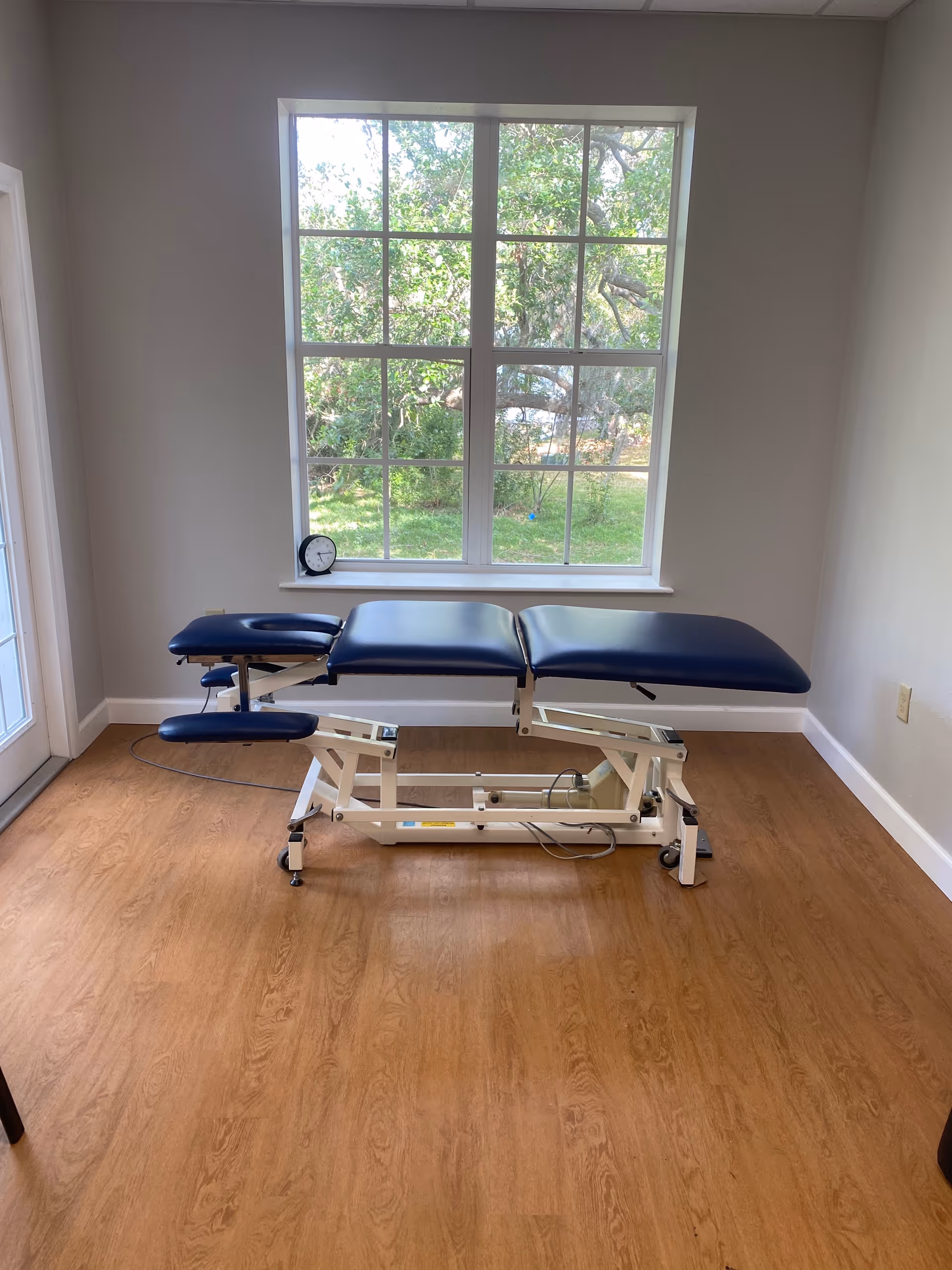Blue padded medical examination table in a bright, empty therapy room with a large window.
