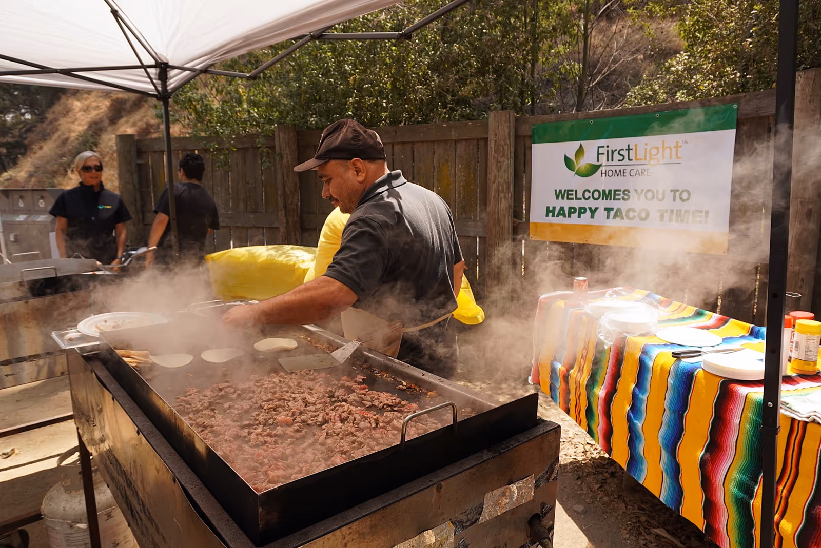 A man grills meat at an outdoor taco event under a FirstLight banner