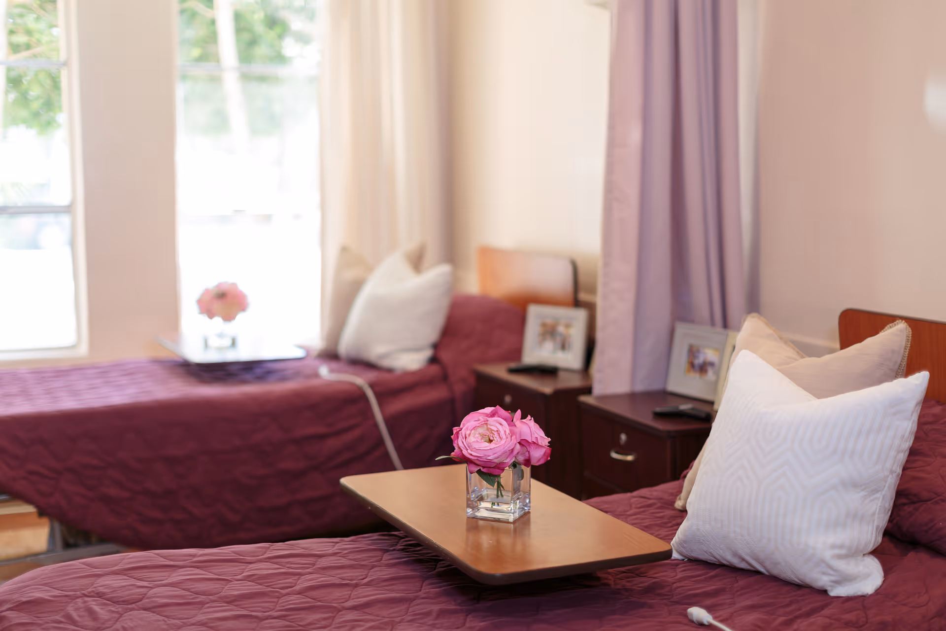 A cozy room with two single beds covered in matching maroon quilts, each bed adorned with white and beige pillows. A small wooden table with a vase of pink flowers is placed between the beds. The room has light-colored walls, a window with sheer curtains letting in natural light, and bedside tables with framed photos and remote controls.