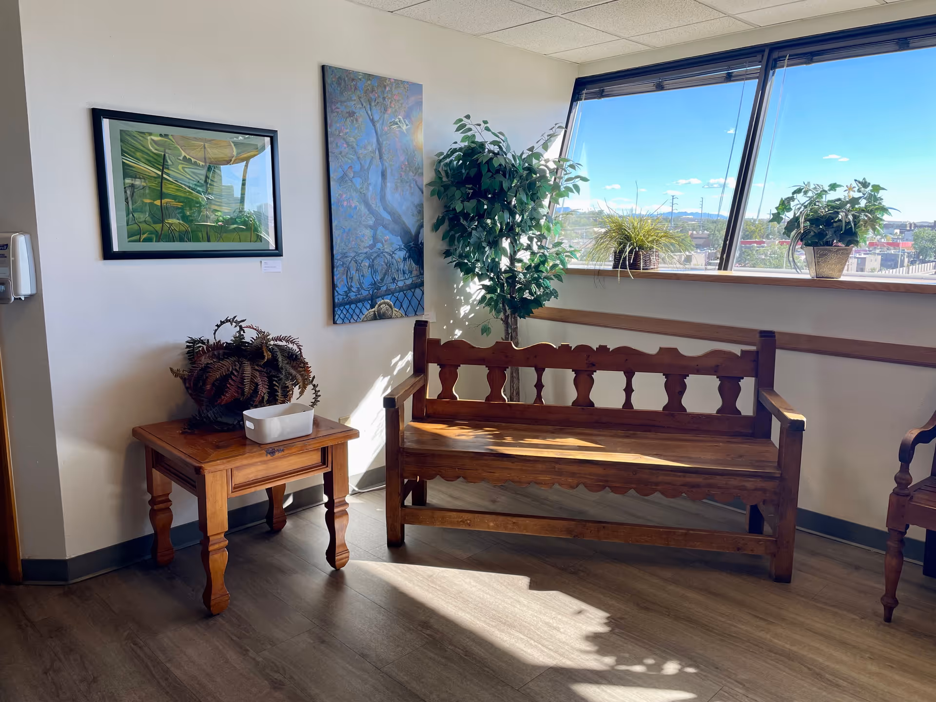 Interior lounge area with wooden bench, end table, plants, artwork, and large windows.