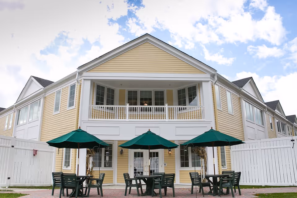 Exterior view of a two-story yellow building with white trim, featuring a balcony on the upper floor and three green patio umbrellas shading outdoor tables and chairs on a brick patio.