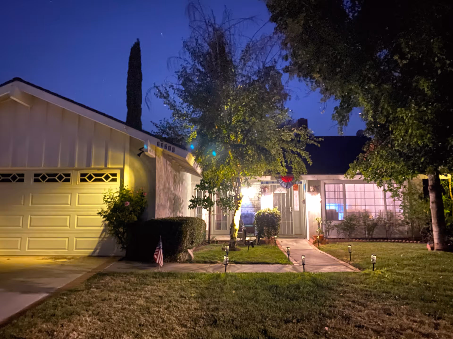A single-story house at dusk with exterior lights illuminating the front yard and entrance. The house has a garage on the left, a walkway lined with small solar lights leading to the front door, and several trees and bushes in the yard. The sky is dark blue indicating early night.