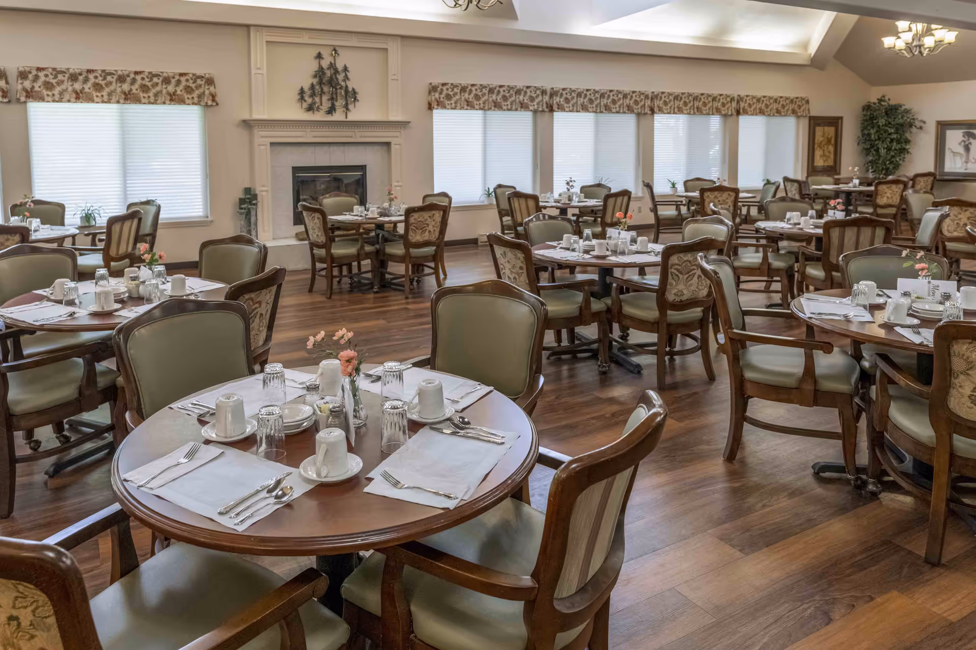 Spacious dining room with multiple round tables set for meals, wooden chairs, and a fireplace along the back wall.