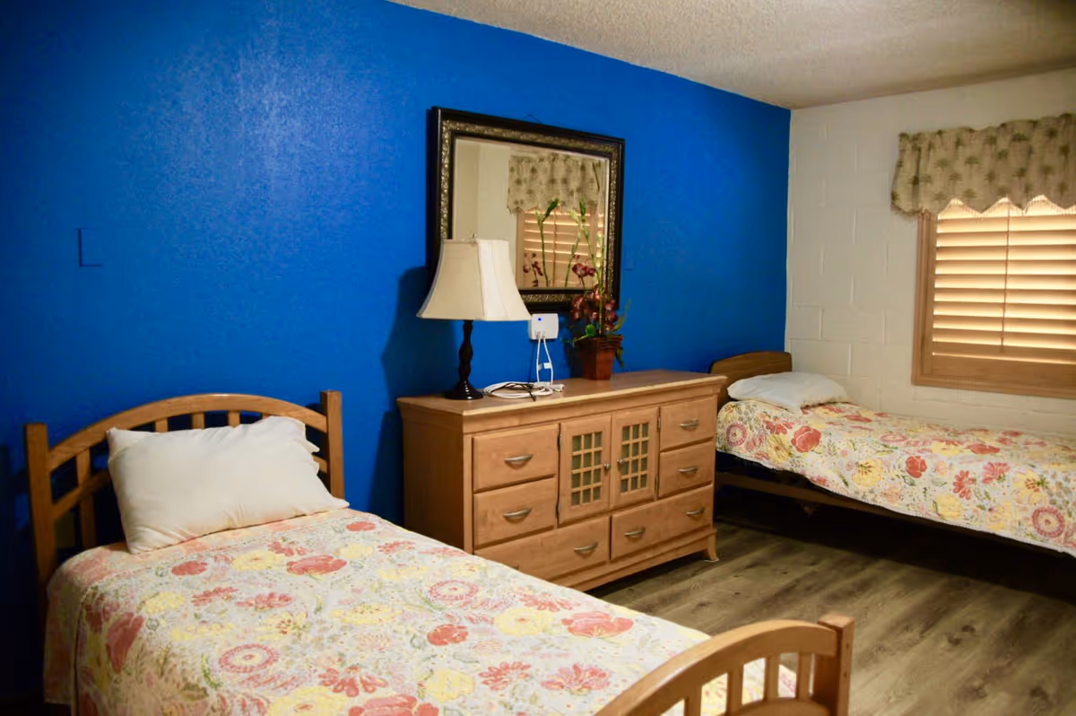 Two single beds with floral covers flank a wooden dresser against a blue wall.