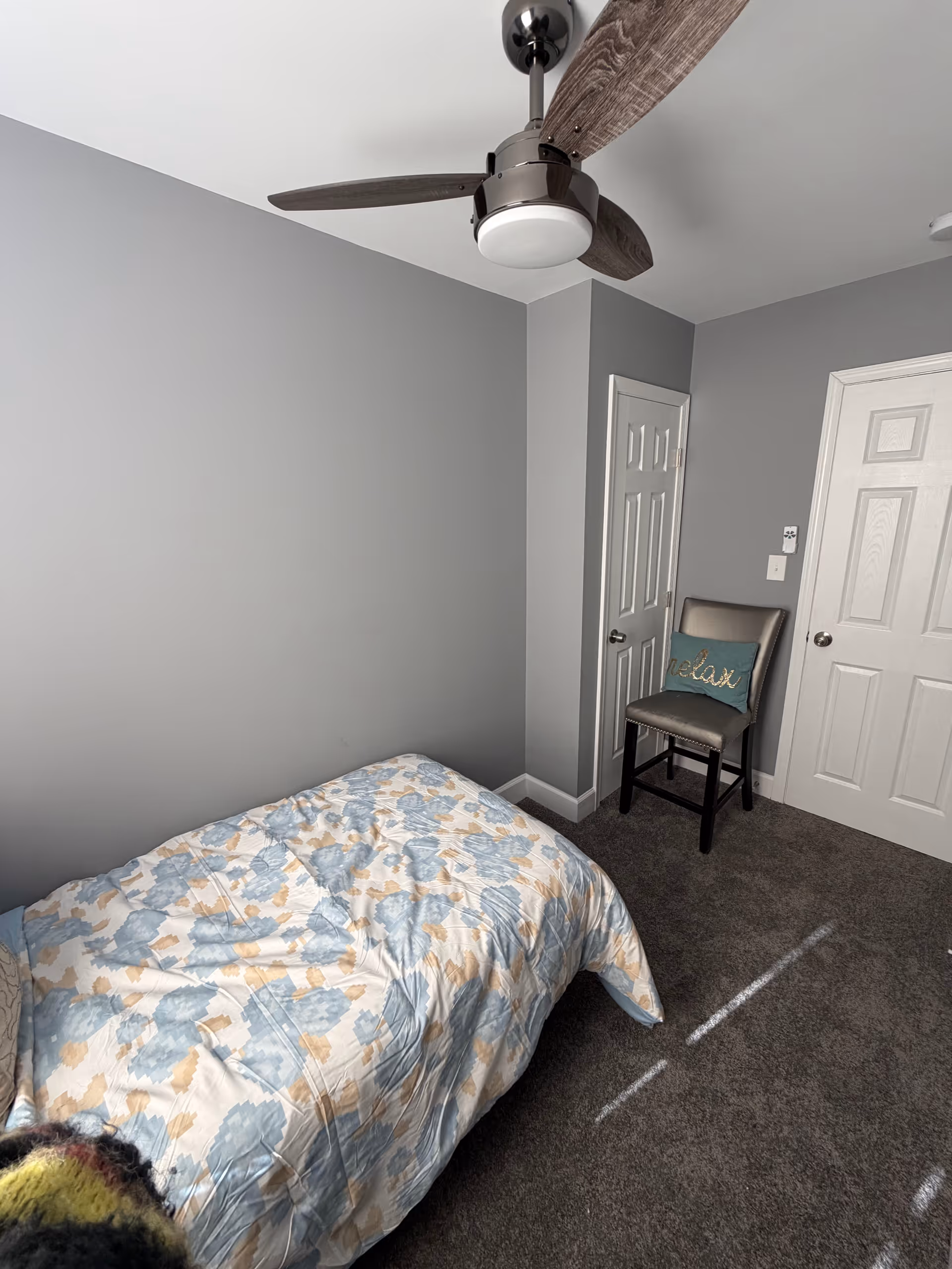 Small bedroom with a floral quilt on the bed, chair, door, and ceiling fan.