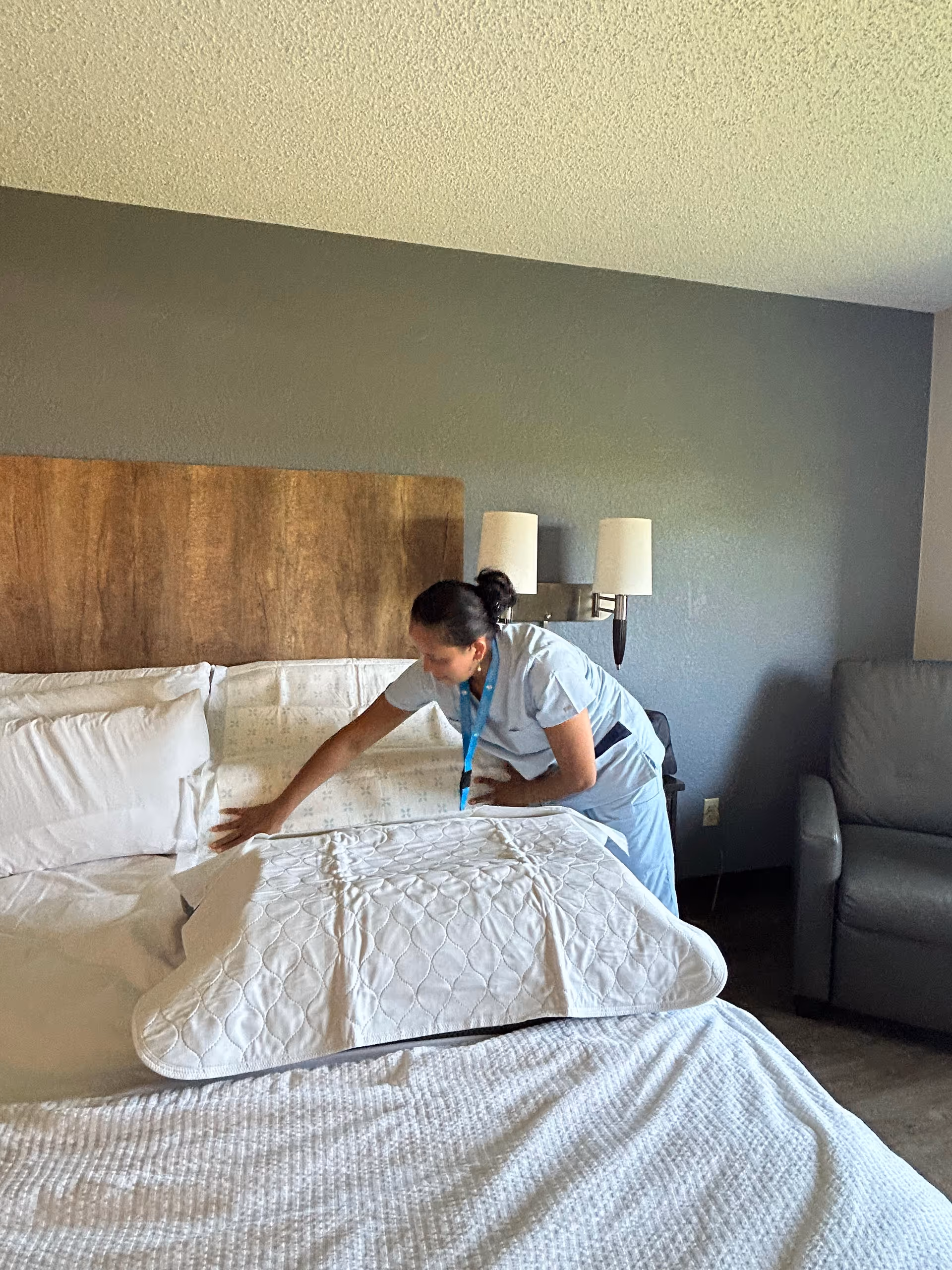 Caregiver in scrubs straightens the bed in a bedroom with a headboard against a gray wall.
