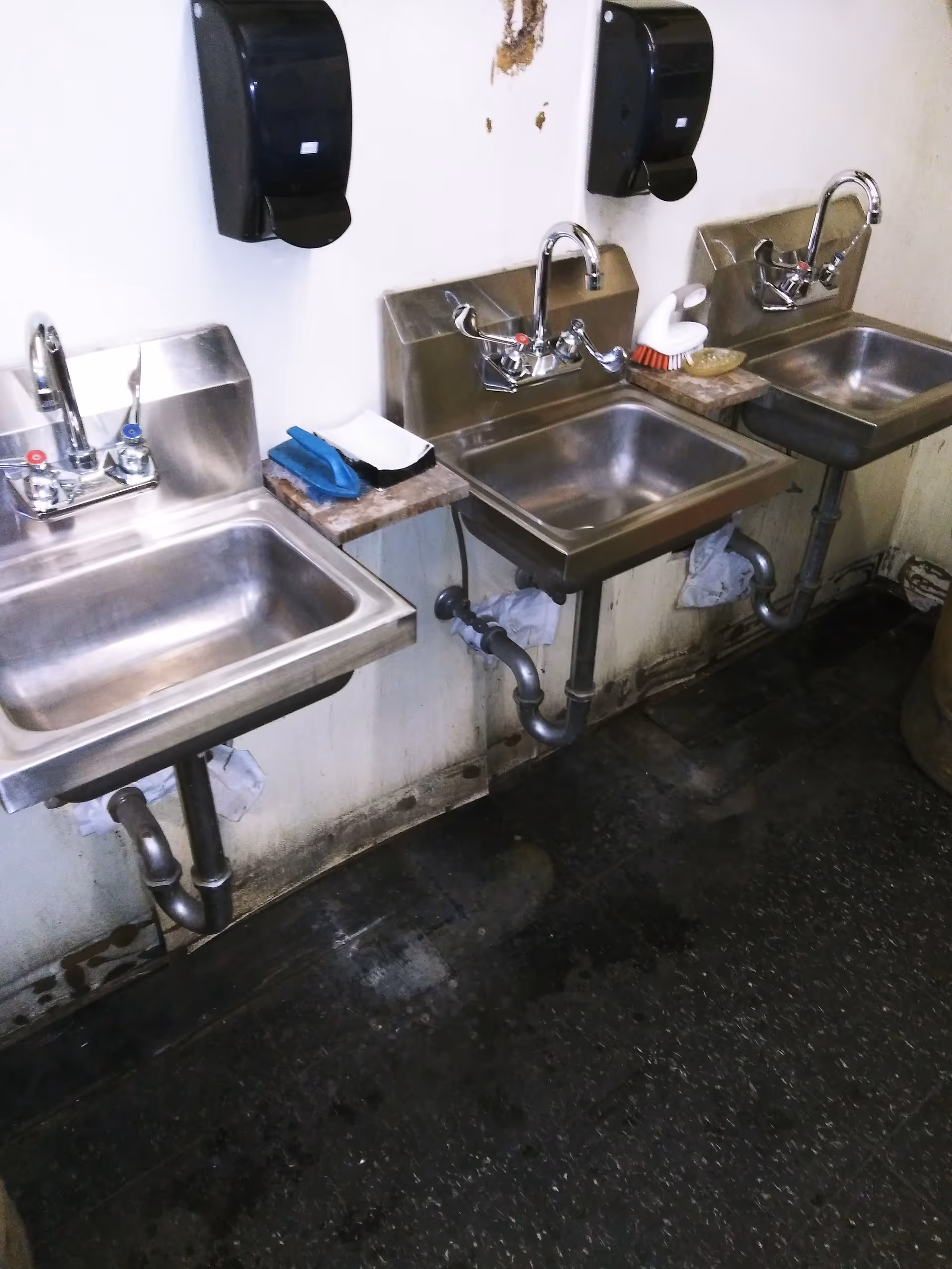 Row of stainless steel sinks with soap dispensers along a dirty utility area.