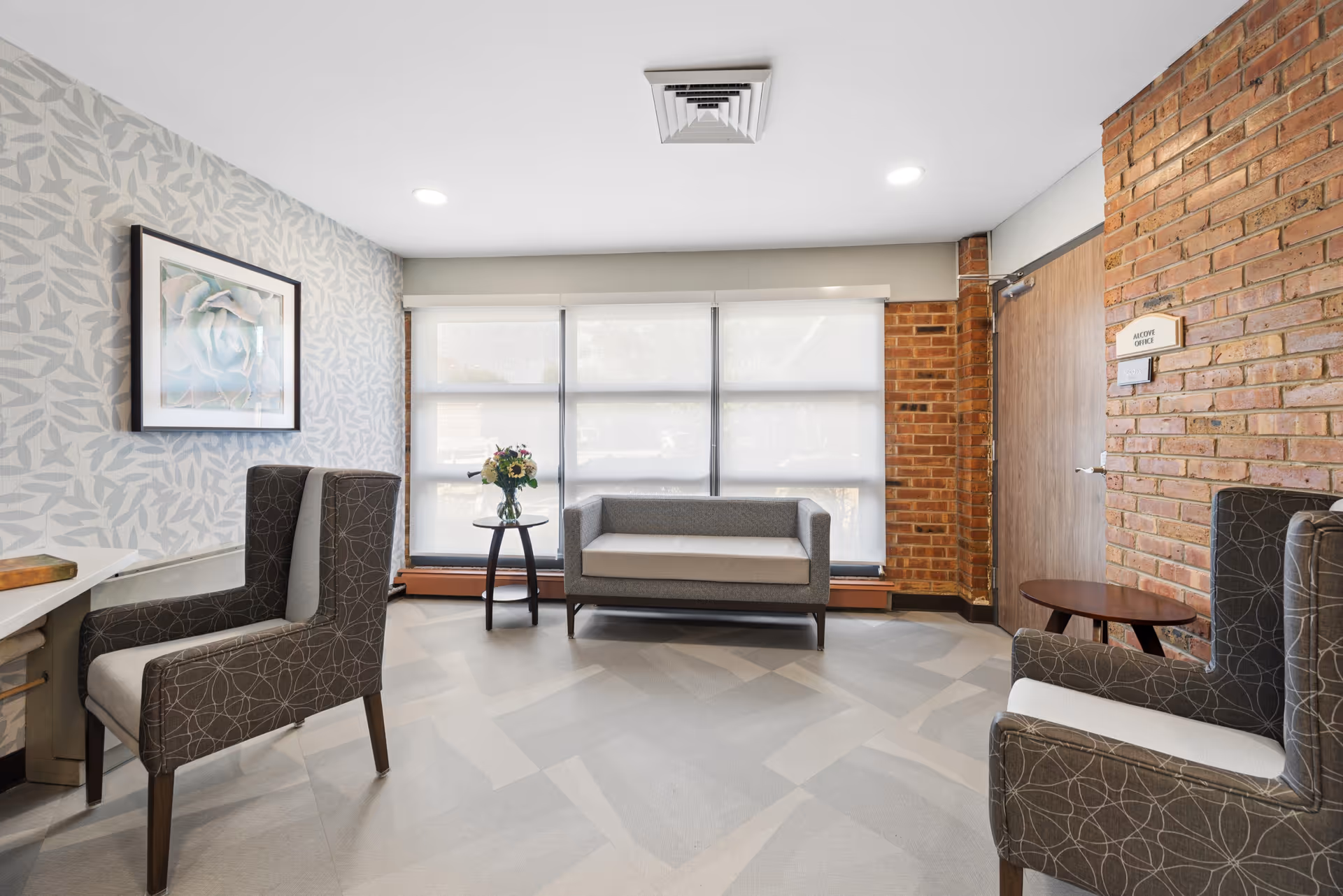 Bright interior waiting area with patterned wallpaper, brick wall, and modern armchairs near windows.