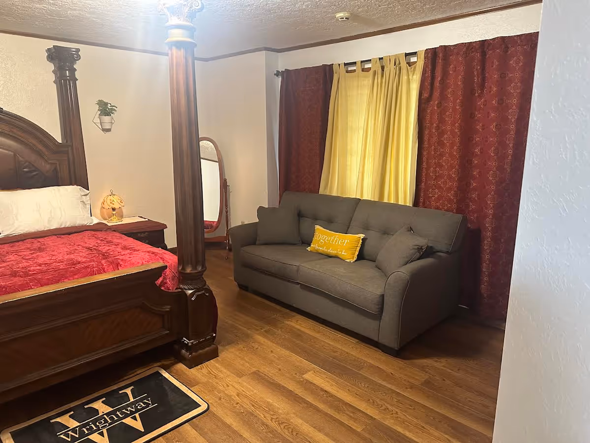 A cozy senior living bedroom with a four-poster bed and a grey sofa under warm curtains.