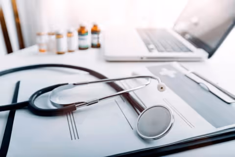 Medical stethoscope on a clipboard with forms, pills, and a laptop.