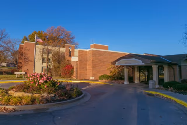Front entrance of a low-rise brick post-acute rehabilitation building with a covered canopy, circular driveway, landscaped island, and an American flag.