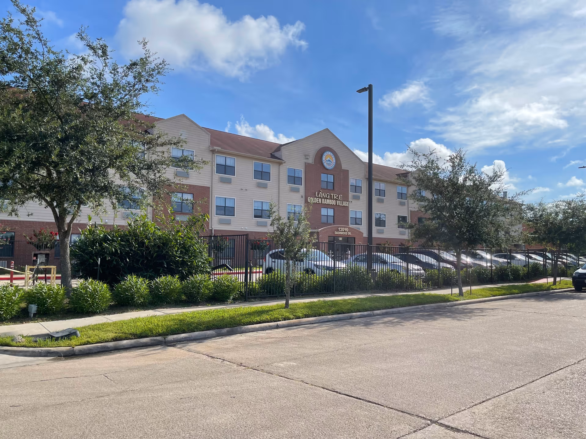 Three-story senior living building with a parking lot, trees, and a black fence.