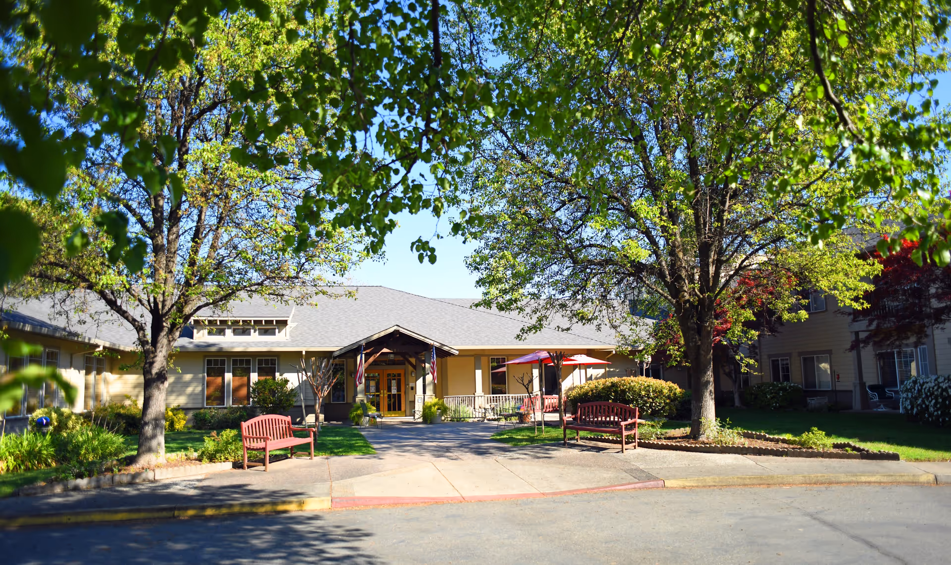 Front entrance of a single-story assisted living building with benches, trees, a paved driveway, and shaded seating.