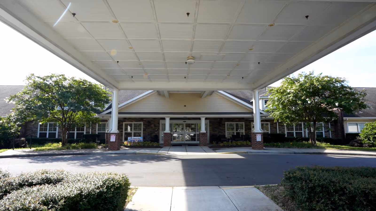 Front exterior view of The Pavilion Health Center at Brightmore showing a covered entrance with white pillars, a driveway, and landscaped bushes and trees on either side.