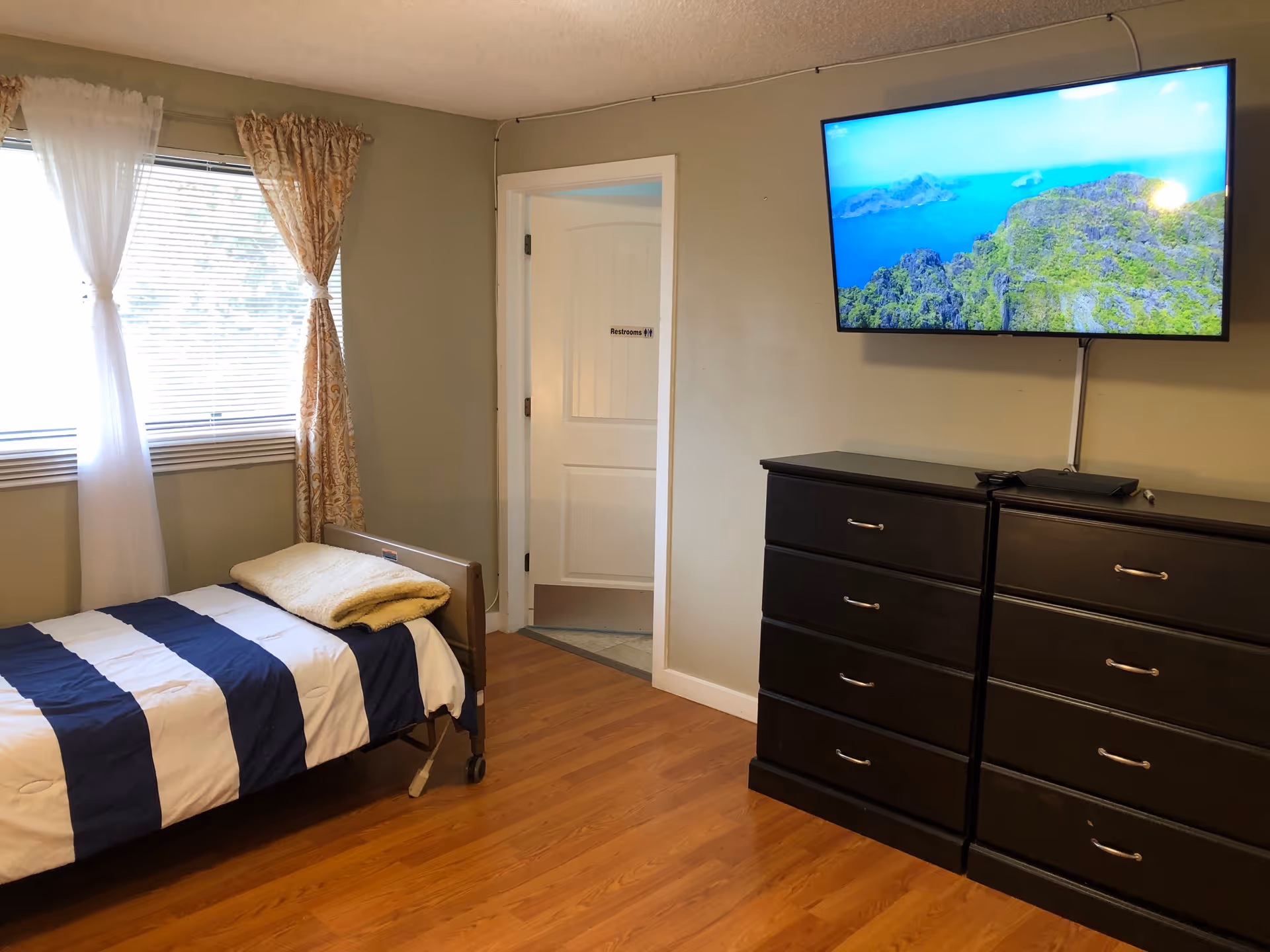 A bedroom with a single striped bed by a window, a wall-mounted TV above two black dressers, and a doorway to a bathroom.