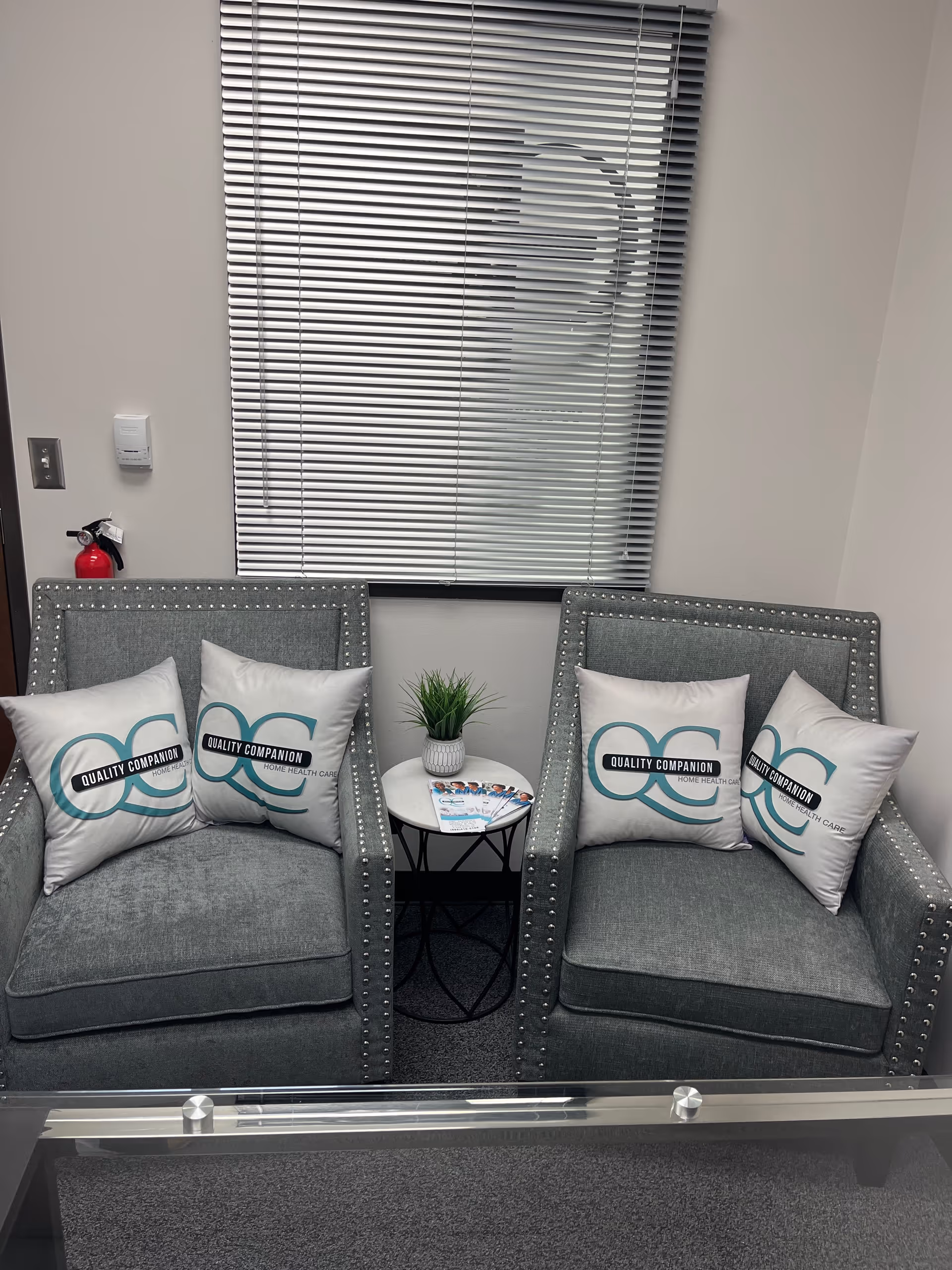 Two grey upholstered armchairs with studded trim flank a small round table and window blinds behind.
