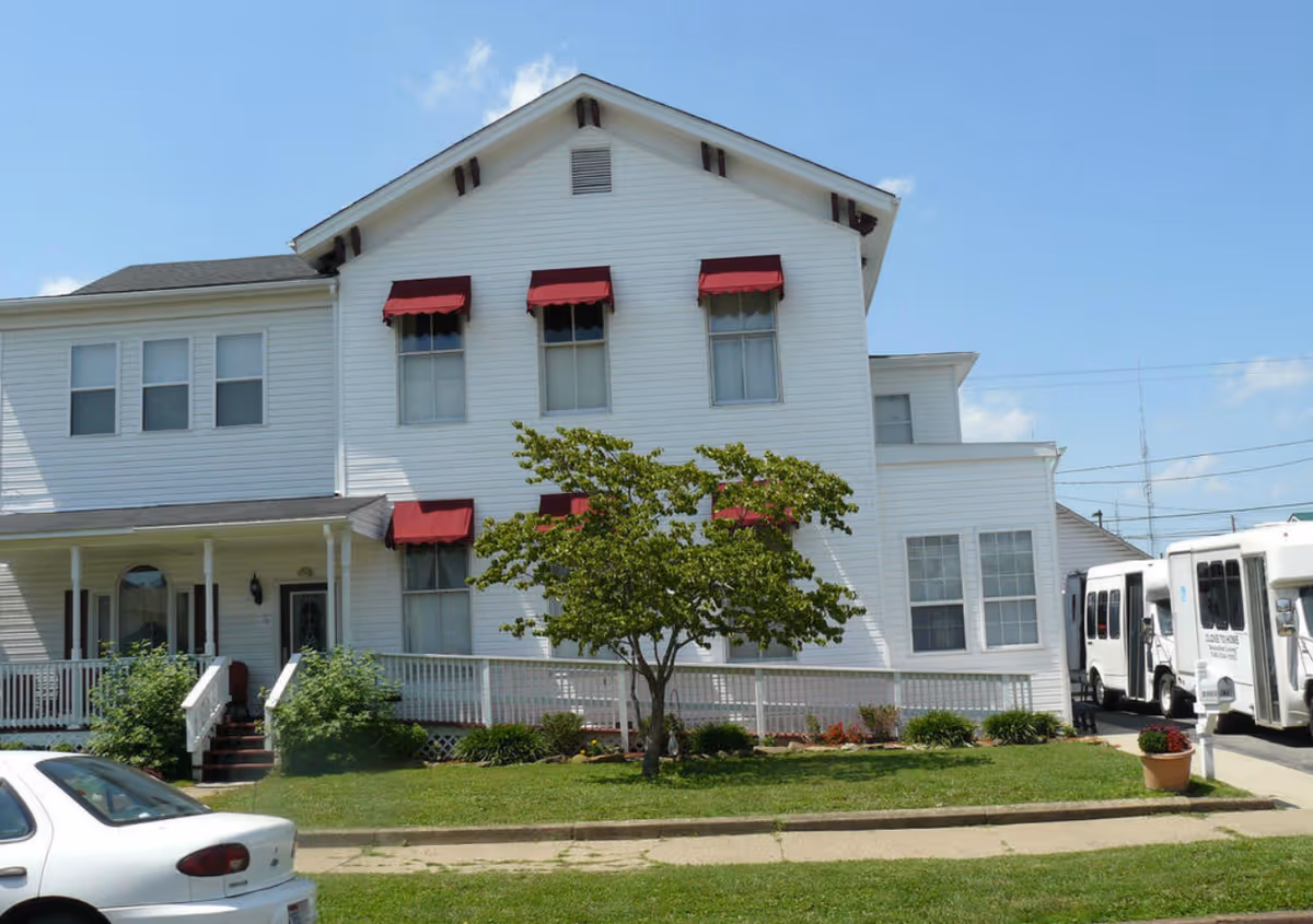 White multi-story senior living facility building with red window awnings, a front porch, small tree and parked vehicles.
