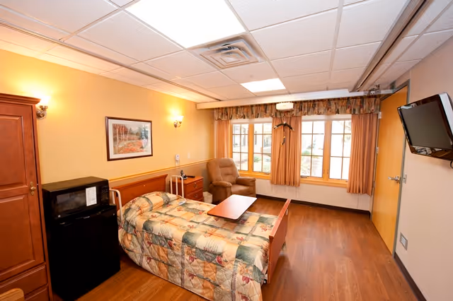 Cozy resident bedroom with quilted bed, chair, desk, and window.
