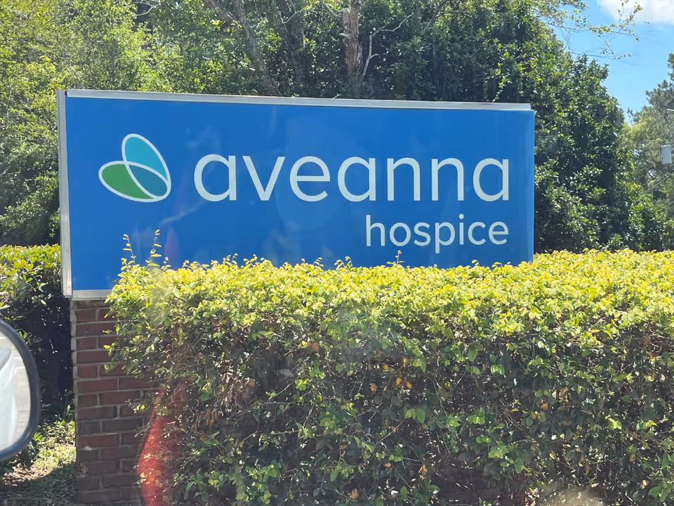 Blue Aveanna Hospice sign mounted on brick base with green hedges outside facility.