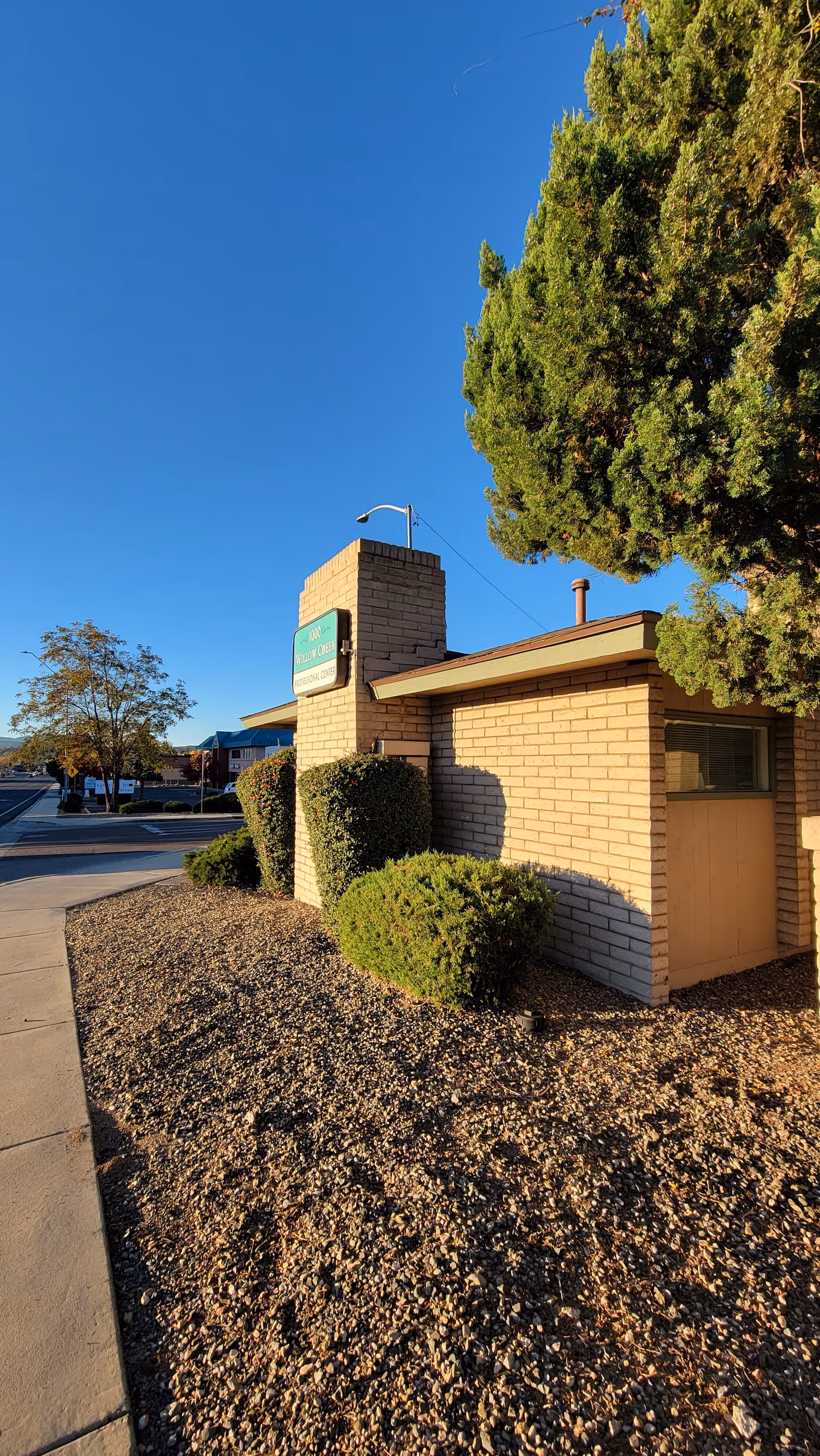Exterior view of Angels Care Hospice building with brick walls, sign, trees, and pebbled landscaping.