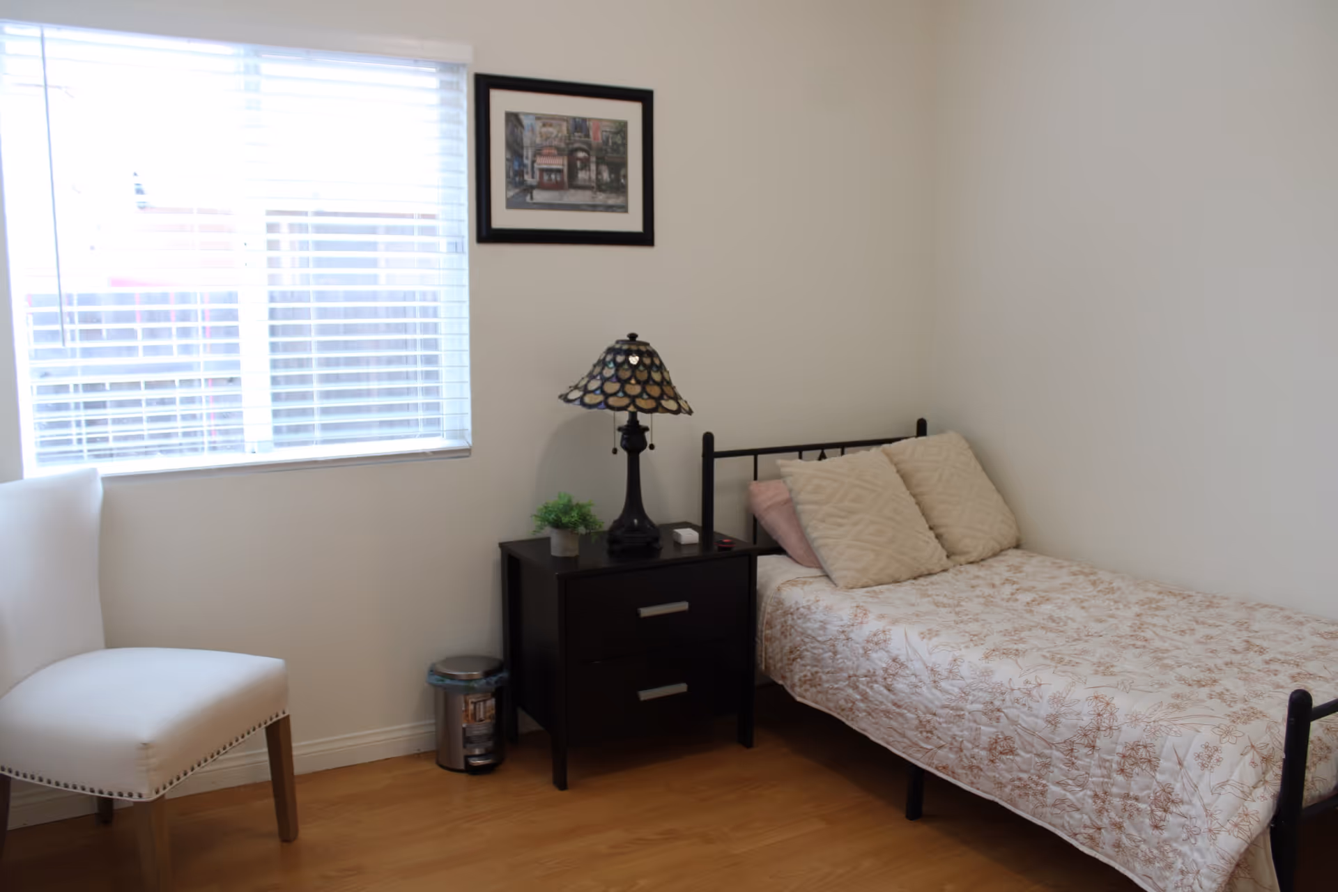 A small, simple bedroom with a single bed covered in a floral-patterned quilt and two beige pillows. Next to the bed is a dark wooden nightstand with two drawers, a decorative lamp, and a small potted plant. A white upholstered chair is positioned near a window with white blinds. A framed picture hangs on the wall above the nightstand. The floor is wooden, and the walls are painted white.