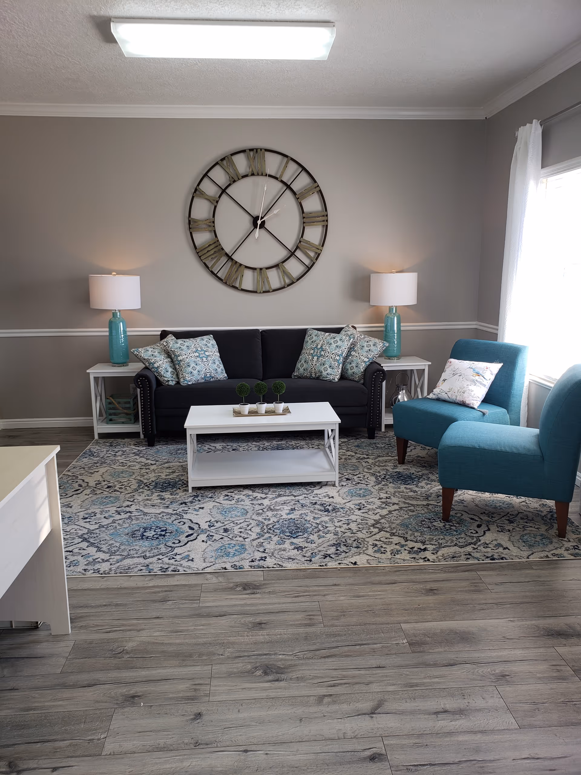 Cozy living room featuring a navy sofa, teal chairs, coffee table, rug, and large wall clock.