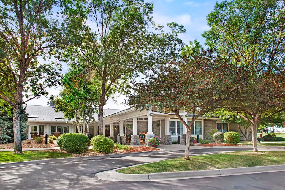 Front entrance of a single-story senior living facility with a covered porch, landscaped shrubs and trees, and a curved driveway.