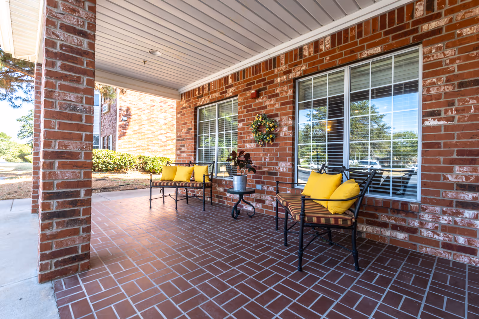 Covered brick front porch with chairs featuring yellow cushions, a small table with a plant, and a wreath on the wall.